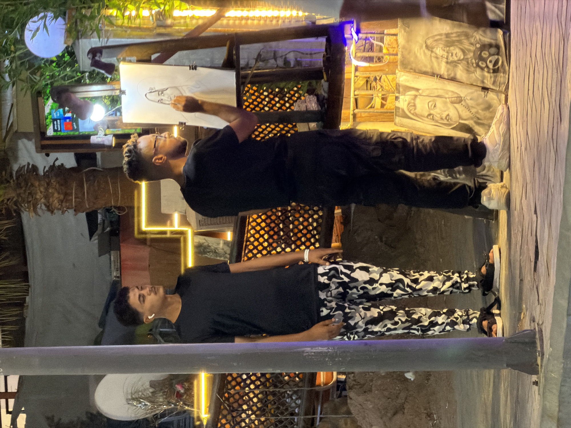 In Sharm El Sheikh, Egypt, at night, a young man with short dark hair and white earbuds stands to the left, wearing a black t-shirt and camouflage pants. He is looking to his right. To his right, another young man with glasses and short dark curly hair, wearing a black t-shirt and black cargo pants, is actively drawing a portrait of a woman on a white canvas on an easel. In front of the easel, to the right, are two charcoal portraits displayed on the ground. One is of Marilyn Monroe, and the other appears to be of a young woman with a teddy bear. The scene is illuminated by warm, yellow string lights and other ambient lighting, creating a vibrant atmosphere. The ground is paved, and a large gray pole stands prominently in the foreground to the left.