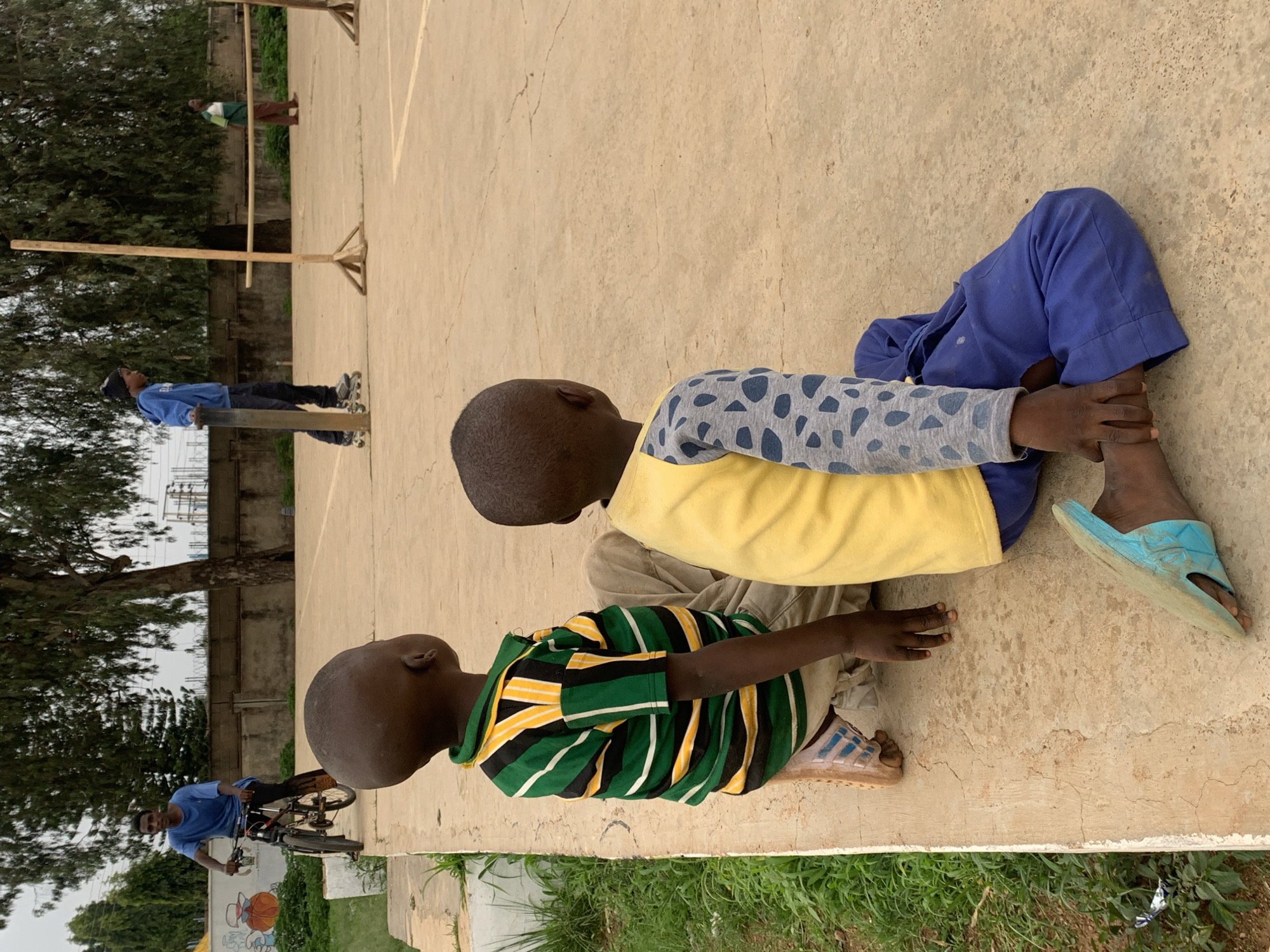 Two young boys are sitting on a concrete surface outdoors. One is wearing a striped green and yellow shirt and light-colored pants. The other is wearing a yellow shirt with gray and blue sleeves and blue pants. The boy in blue is touching the other boy's leg. The location appears to be a public space or schoolyard, evidenced by the presence of other individuals in the background: one riding a bicycle and others by wooden structures. The environment includes trees and vegetation. It is daytime. The scene is located in Jos, Nigeria.