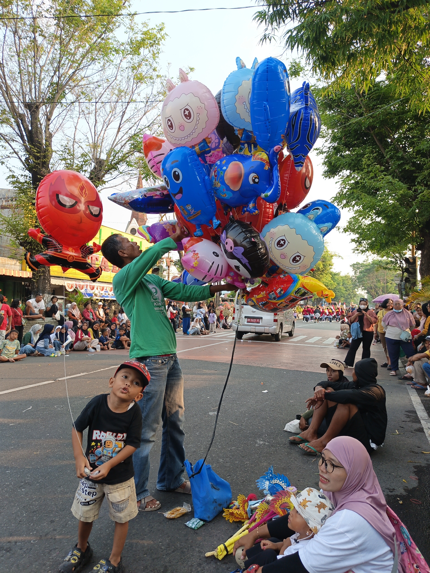 In Klaten, Indonesia, a balloon vendor, a man in a green long-sleeved shirt and blue jeans, is holding a large bunch of colorful balloons. The balloons are shaped like various characters, including Spiderman, an elephant, a cat, and a fish. In the foreground, a young boy in a black t-shirt and khaki shorts looks up at the balloons. Several other people are sitting on the side of the road, watching the scene. The environment is an urban street with trees lining the road and buildings in the background. The image appears to be taken during the daytime, with the sun casting shadows on the ground.
