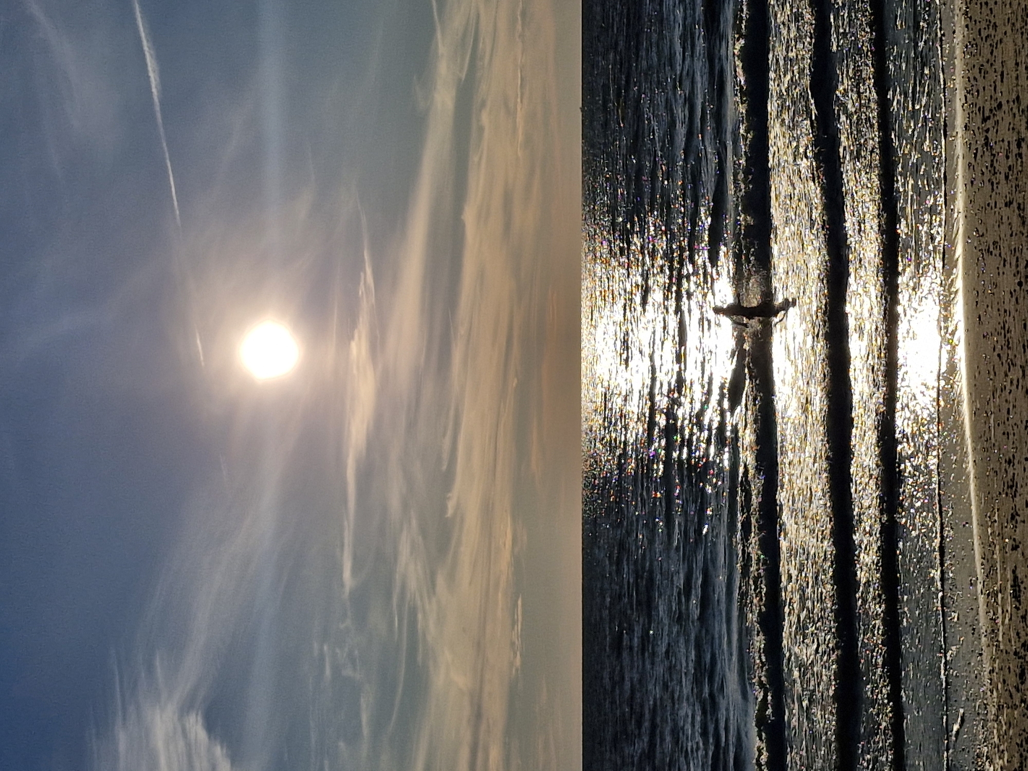 A lone figure stands in the shallow waters of the Baltic Sea at sunset, near the village of Karklė, Lithuania. The sun, low on the horizon, casts a bright, shimmering reflection across the rippling water, creating a path of light that leads to the figure. Wispy clouds streak across the pale blue sky, tinged with the warm hues of the setting sun. The beach in the foreground is a sandy expanse dotted with small pebbles. The overall scene evokes a sense of tranquility and solitude.