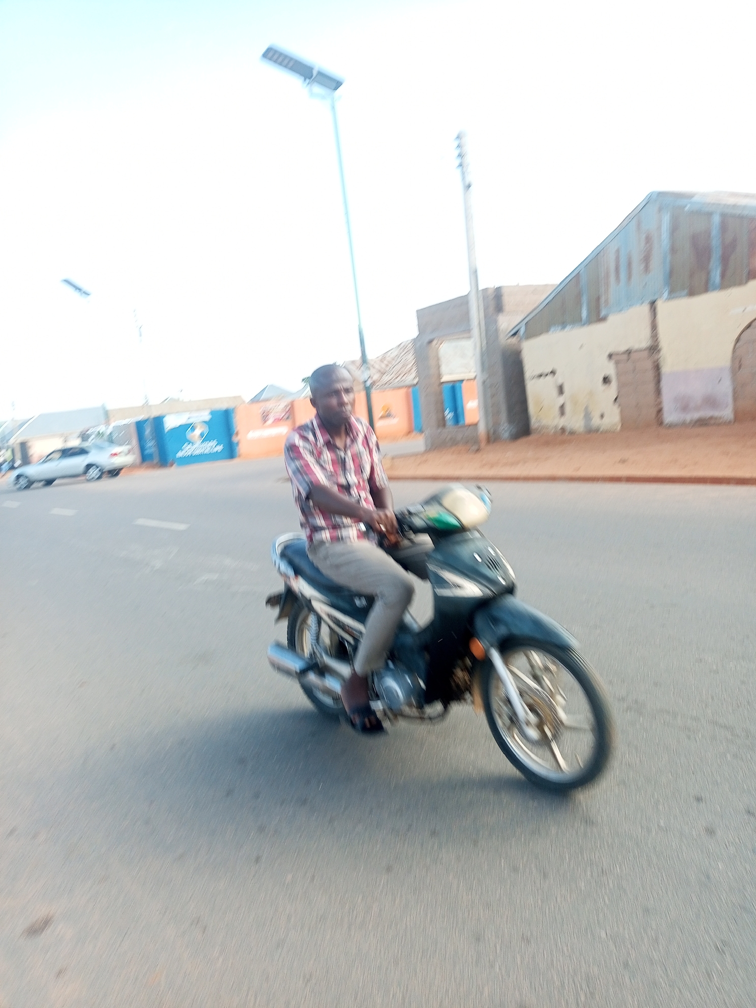 A man is riding a motorcycle on a paved road in Tempure, Nigeria. He is wearing a plaid shirt and light-colored pants. The motorcycle is dark-colored with silver accents. The road is mostly empty, with a few buildings visible in the background. The buildings appear to be made of brick and metal, and some have corrugated metal roofs. There is a street lamp in the distance, suggesting it is either early morning or late evening. The sky is bright and clear, indicating good weather. The image is somewhat blurry due to motion, suggesting the photo was taken while the motorcycle was moving. The overall scene depicts a typical day in a Nigerian town. No visible text is present in the image.
