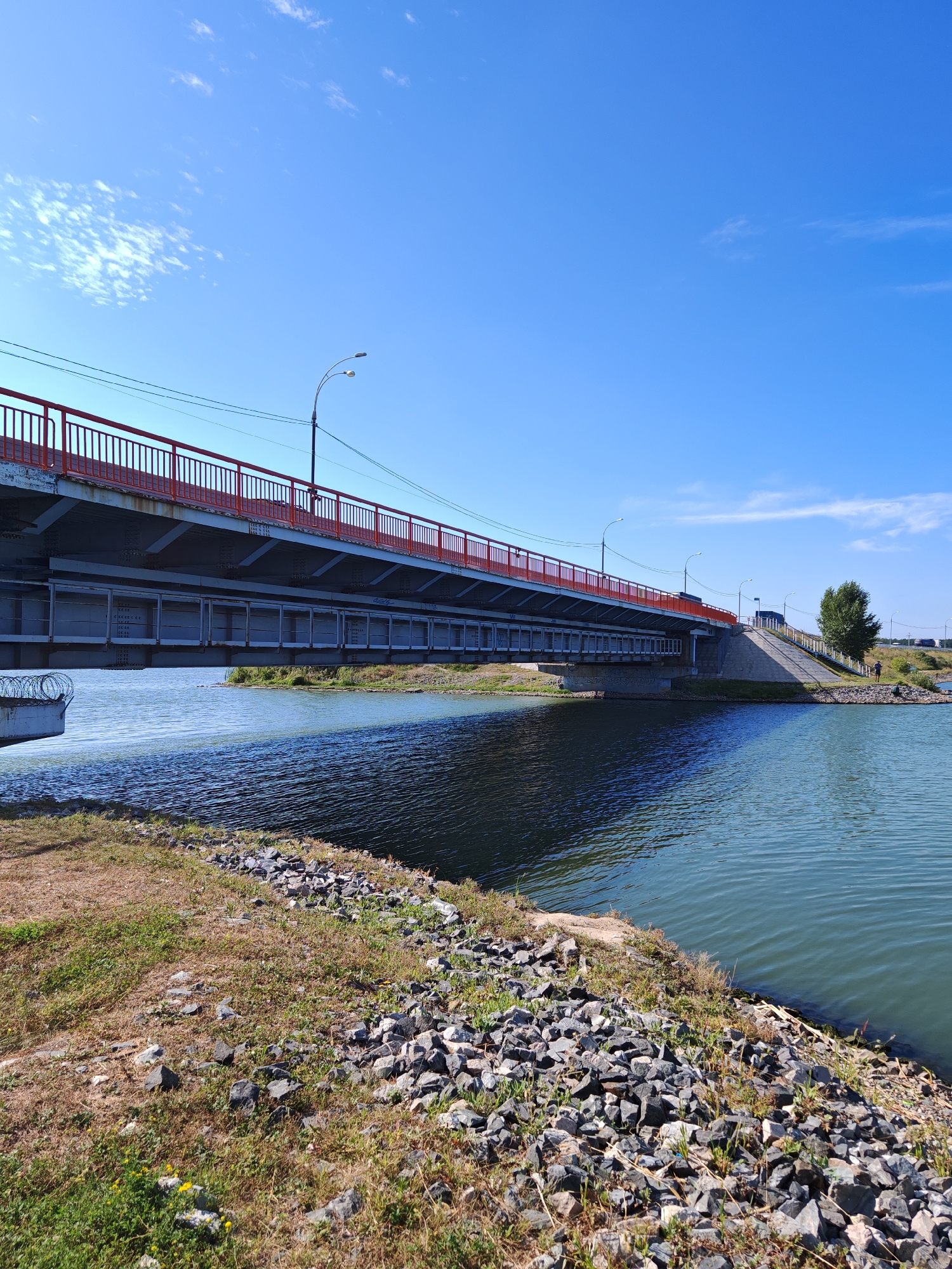 A clear, sunny day with a bright blue sky and scattered white clouds over the city of Chernakovo, Russia. A modern bridge with red railings spans a calm body of water. The bridge's structure is visible underneath, with steel supports and rectangular panels. The water reflects the blue sky and the underside of the bridge. The foreground features a grassy embankment with scattered rocks and some dry vegetation. On the right side of the image, a ramp leads up to the bridge, with a single person standing near a tree. Another person is visible further down the road beyond the bridge. The scene suggests a peaceful, everyday setting by the water.