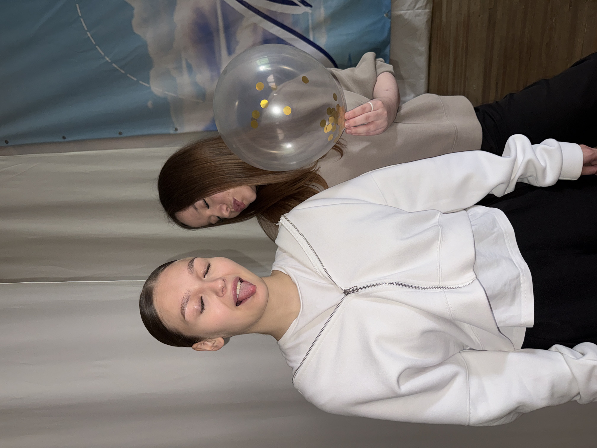 Two young women are posing for a picture indoors. The woman on the left, with her hair pulled back, is sticking out her tongue. She is wearing a white zip-up hoodie over a white t-shirt and black pants. The woman on the right, with long brown hair, is puckering her lips and holding a clear balloon filled with gold confetti. She is wearing a beige sweatshirt and black pants. They are standing in front of a light-colored curtain and a blue banner with a cloud design. The floor is made of wooden planks. The location is the city of Chaikovsky, Russia.