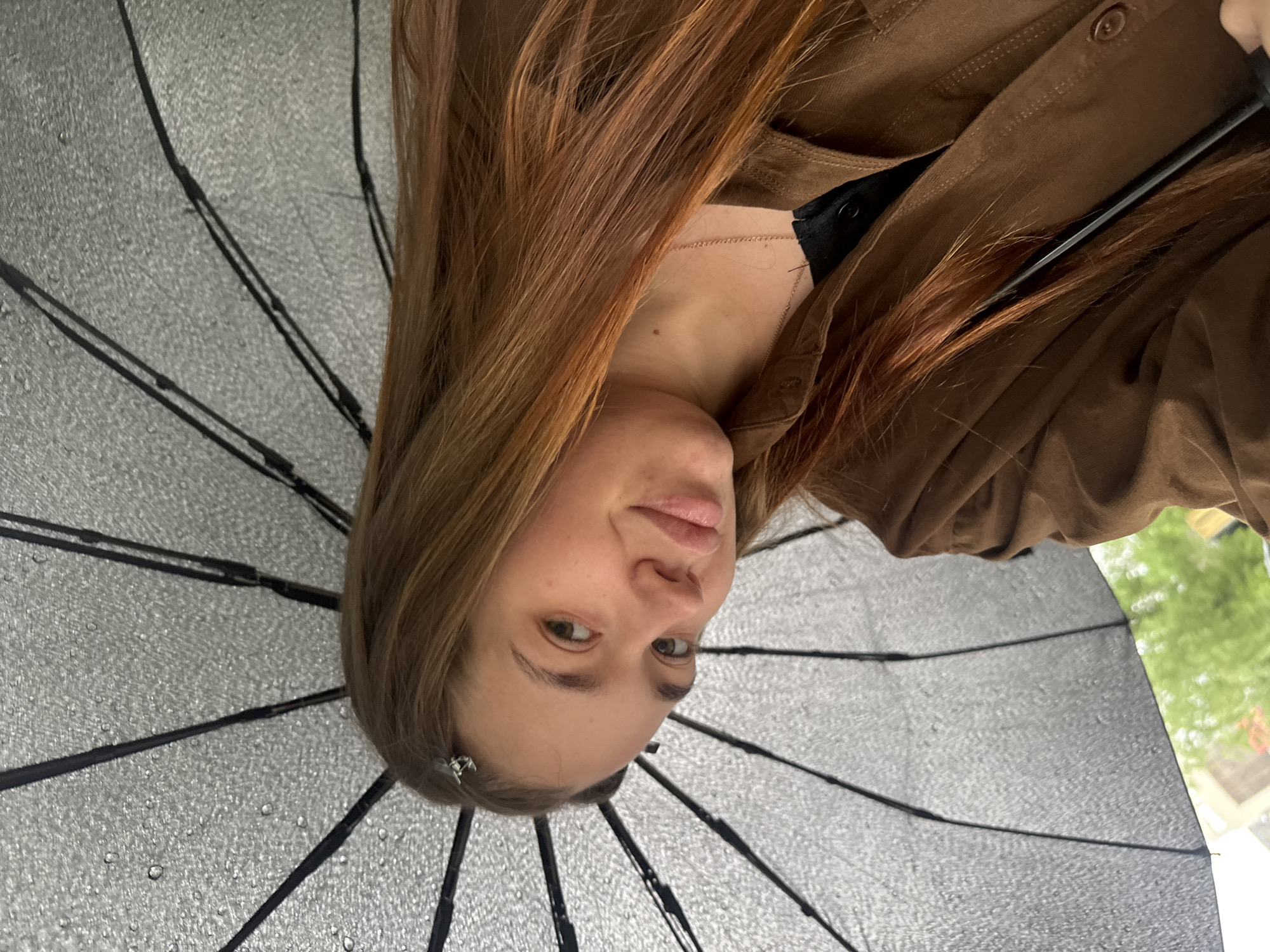 A young woman is pictured from a low-angle, slightly off-center perspective. She is holding a dark-colored umbrella. Water droplets are visible on the umbrella's surface. The woman has light skin, brown hair, and is wearing a brown jacket over a black top. A thin, gold necklace is visible around her neck. Outside the umbrella, there is a glimpse of green foliage, possibly trees. The location is Chaikovsky, Russia.