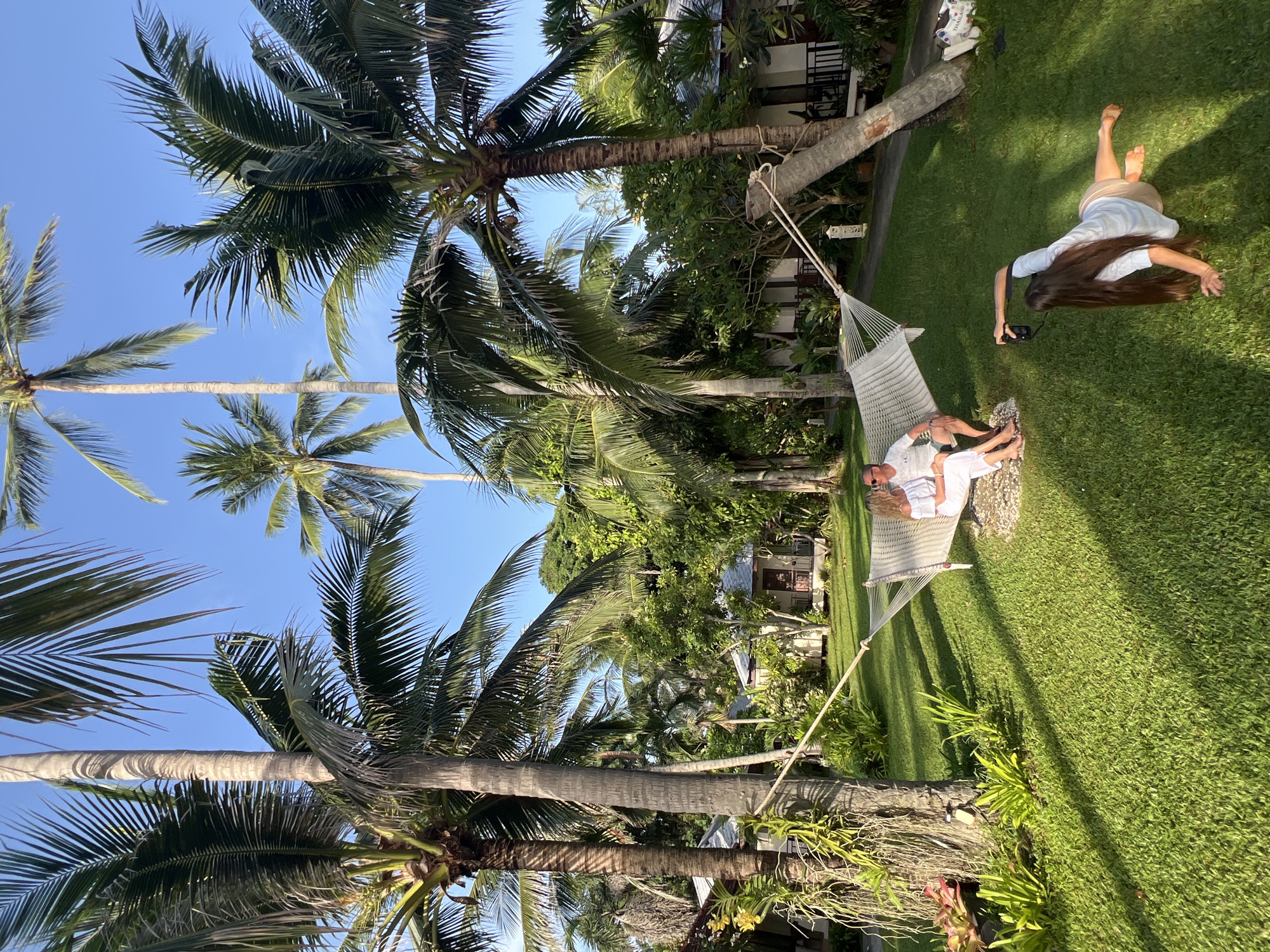 The image captures a serene scene in the Ko Samui District of Thailand, characterized by lush tropical vegetation and clear blue skies. In the foreground, a hammock is strung between two palm trees, supporting a couple dressed in white, seated comfortably and appearing to relax. Beside them, a woman with long dark hair lies on the grass, holding a camera and seemingly taking a photograph. The background reveals several small resort-style buildings nestled amongst dense greenery and more palm trees, suggesting a tranquil vacation setting. The sun casts long shadows across the vibrant green lawn, indicating it's likely late afternoon. The overall ambiance is one of leisure and idyllic tropical escape.