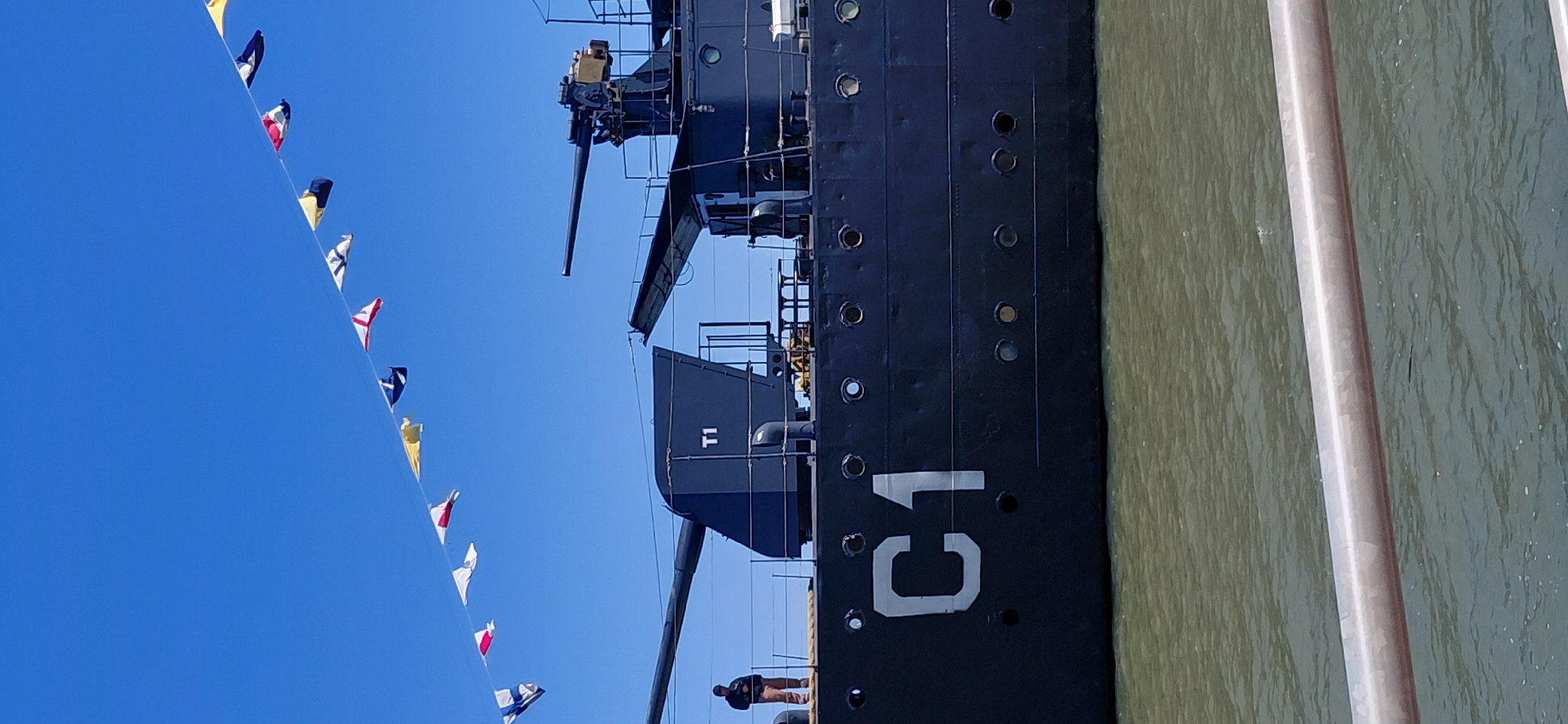 The image depicts the side of a dark-colored ship, possibly a museum ship, docked in the port of Encarnacion, Paraguay. The ship has the designation "C1" painted in large white letters on its hull. A person, wearing a dark top and light-colored pants, stands on the deck of the ship, looking towards the left. The ship is equipped with at least two large gun turrets. In the background, a string of colorful signal flags flies against a clear blue sky. The water in the foreground appears choppy and green. The scene suggests a sunny day, with the ship serving as a focal point against the backdrop of the sky and water.