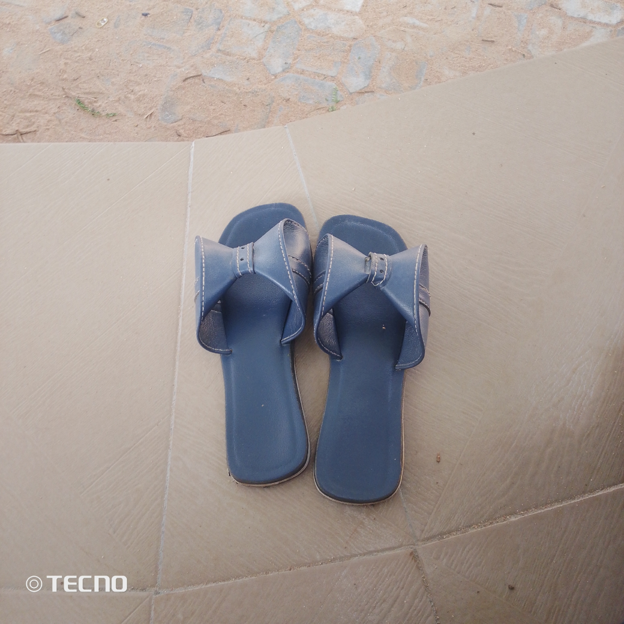 A pair of dark blue, bow-adorned slide sandals are resting on a beige tiled surface. The sandals are positioned side-by-side, with their soles facing upwards. The background behind the tiled area transitions to a sandy surface with scattered small stones and what appears to be a cobblestone path. A watermark with the TECNO logo is visible in the bottom left corner of the image. The overall setting suggests an outdoor area, possibly a patio or walkway, in Jalingo, Nigeria.