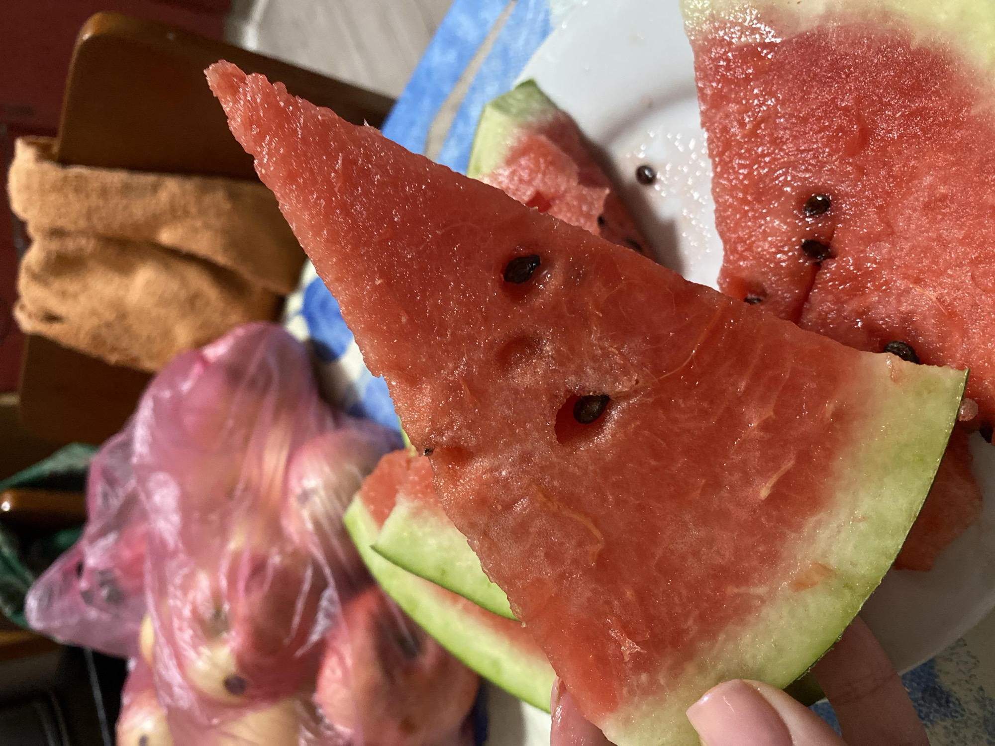 A close-up shot of a person holding a slice of watermelon over a plate of other watermelon slices. The watermelon is bright red with visible black seeds. In the background, there are pink plastic bags filled with fruit, a folded orange towel, and a wooden chair. The setting appears to be a home environment, possibly a kitchen or dining area. The exact location in Chornomorsk, Ukraine cannot be determined from the visual information provided.
