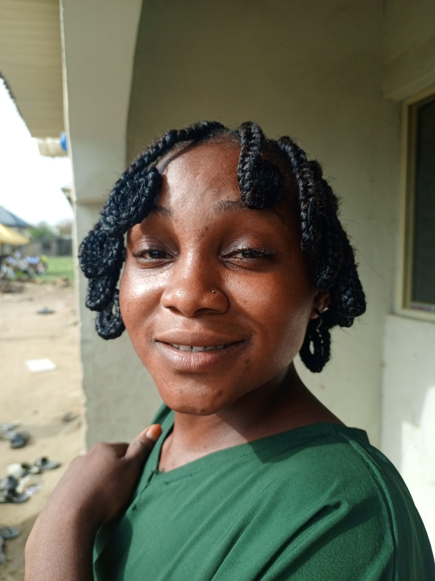 The image is a close-up shot of a young Black woman with dark, styled braids framing her face. She is wearing a green shirt and smiling slightly. She has a nose piercing. The background features an outdoor setting, possibly a residential area, with hints of buildings and other people visible in the distance. The image appears to have been taken in natural daylight. The specific location in Keffi, Nigeria is not explicitly identifiable from the given image.