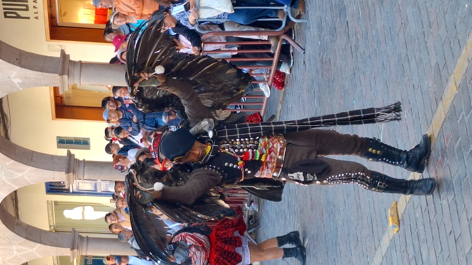 This image captures a vibrant street scene in Cuzco, Peru, likely near the Plaza de Armas, with its characteristic colonial architecture. A performer, dressed in a striking costume that includes a black hat, a dark vest adorned with silver studs and colorful tassels, and brown pants, stands center frame. The performer is holding large, dark feathered wings, outstretched to either side, resembling those of a bird of prey. Their face is partially obscured by a black mask or headdress. The performer wears tall, black, studded boots.
To the left of the performer, a woman in a red, layered skirt and white undergarment is visible. A crowd of onlookers stands behind a metal barrier, observing the performance. The ground is paved with cobblestones. In the background, arched walkways and buildings with signs are discernible, suggesting a commercial area. The lighting indicates it is daytime.