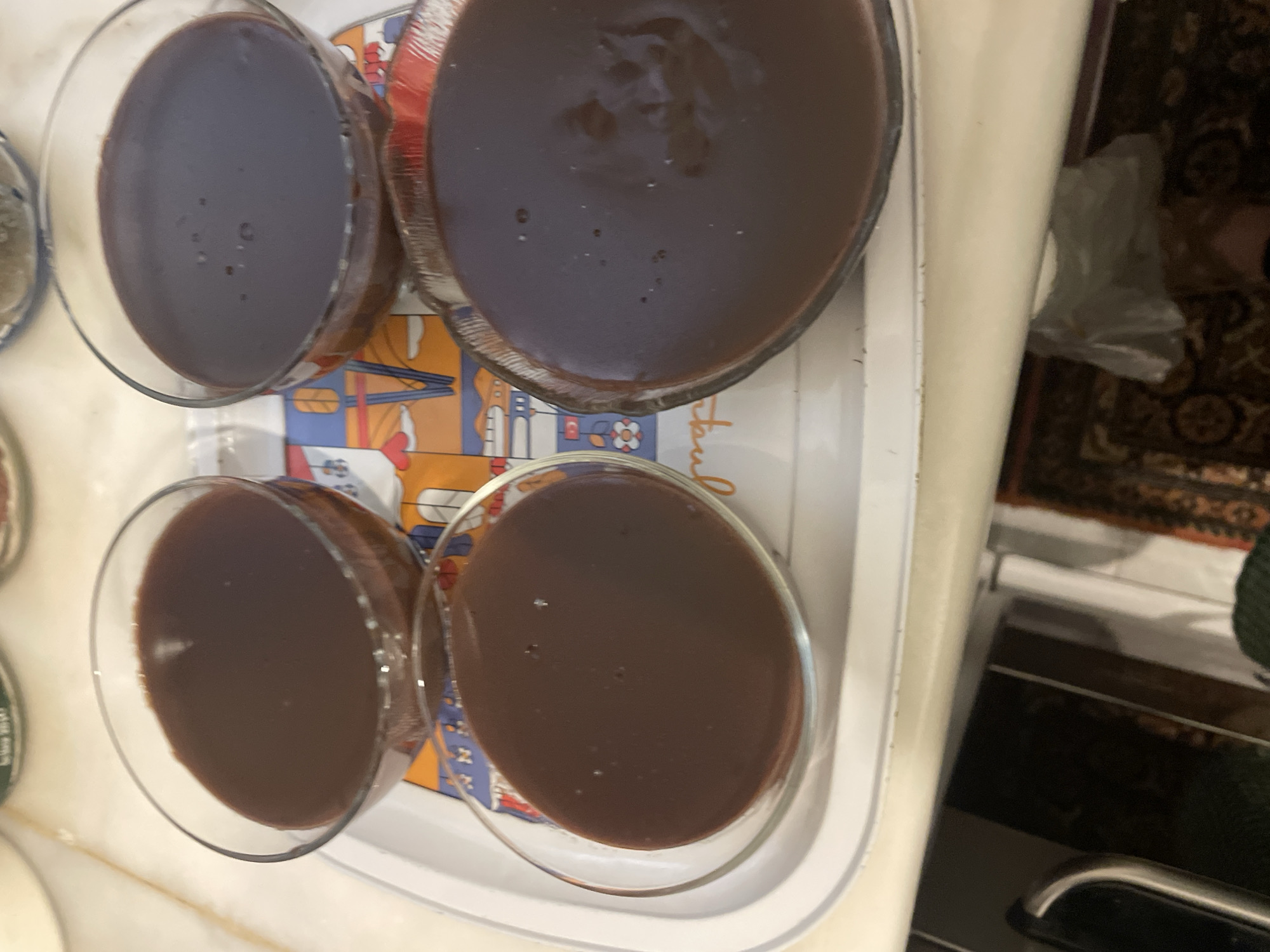 The image shows four glass bowls filled with a dark brown, semi-liquid substance, likely chocolate pudding or mousse. The bowls are arranged on a white tray with colorful illustrations on its sides. The tray is placed on a surface that appears to be a table. The background is blurred, but suggests an indoor setting with a patterned rug visible below the table. There are no people visible in the image, and no specific location in Kadıköy, Turkey, can be identified from the visual information provided. The lighting is overhead and appears to be artificial, casting soft shadows.