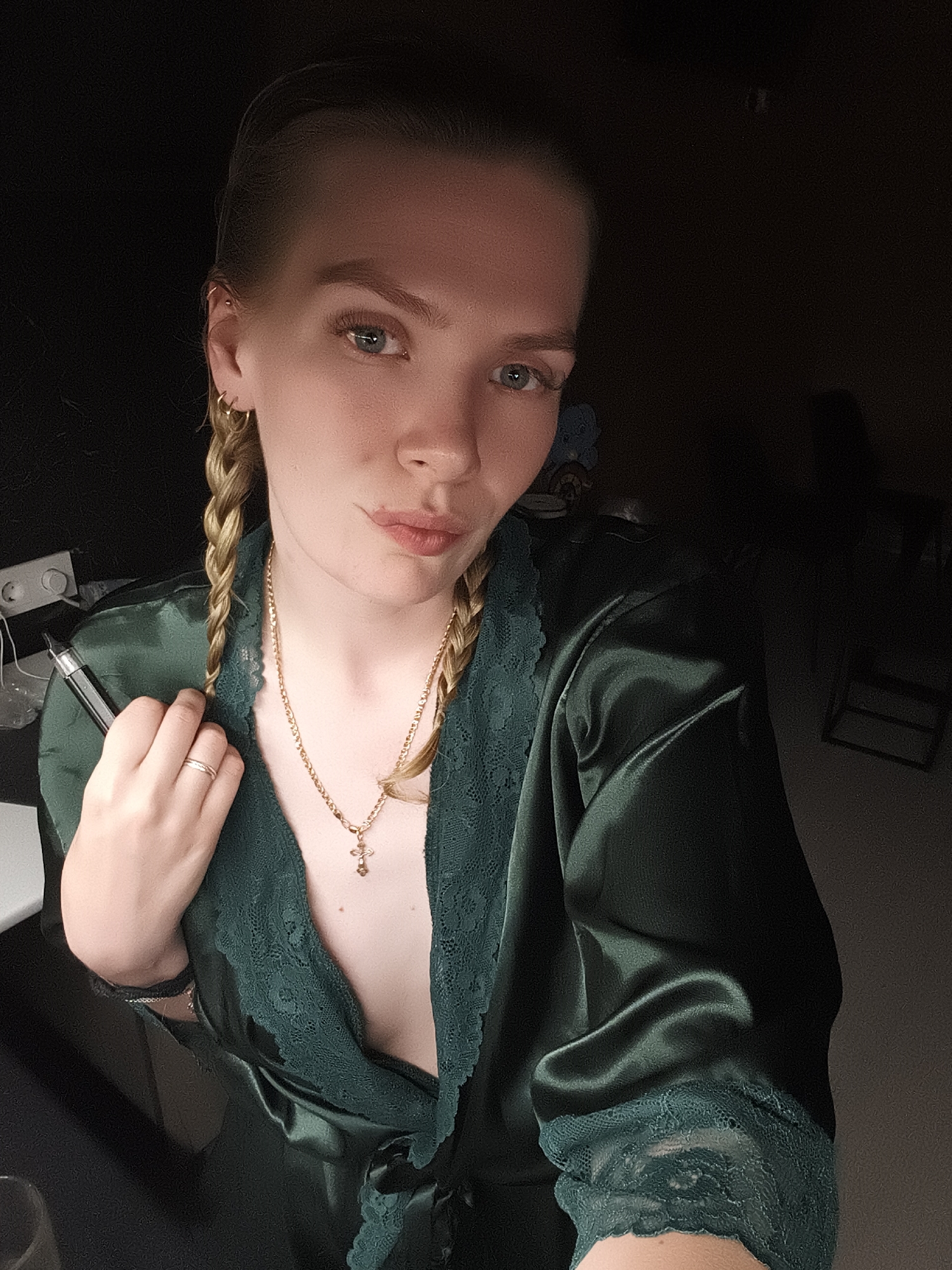 The image is a medium shot selfie of a fair-skinned woman with blonde hair styled into two braids. She is wearing a deep green satin robe with lace detailing around the neckline and sleeves. She is holding a black electronic cigarette in her right hand. A gold necklace with a cross pendant is visible around her neck. She has a neutral expression with a slight pout. Her eyes are light blue. She has multiple piercings in her left ear. A gold ring is on her right ring finger.
In the blurred background, a white electrical outlet is visible on a dark surface to the left. Behind her, there is a dark room, and a chair can be faintly discerned.
There is no information to ascertain this photo being taken in Русский Вожой, Russia.