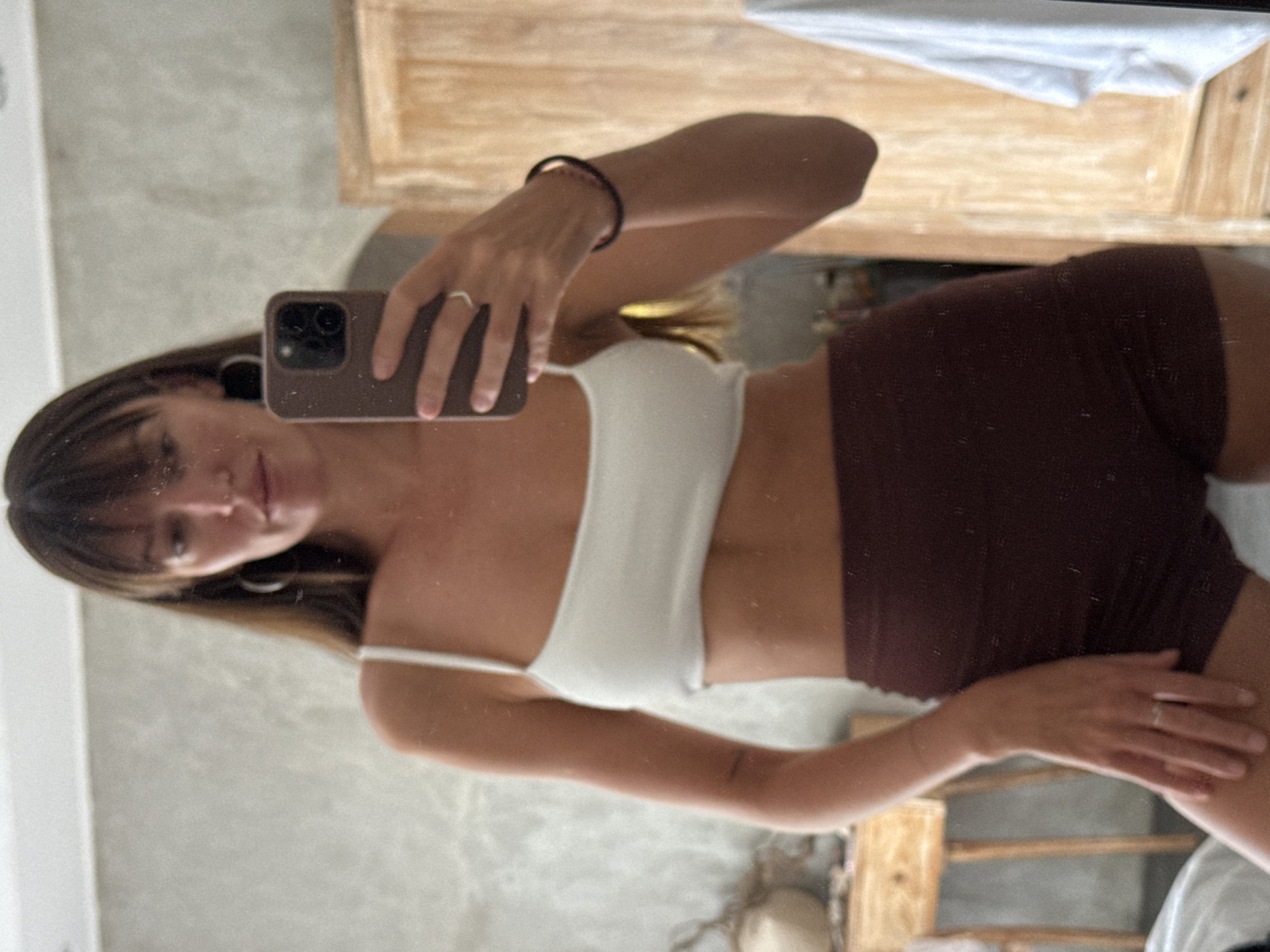 A full-body mirror selfie shows a woman with long brown hair and bangs, wearing a white spaghetti strap crop top and dark brown high-waisted shorts. She has hoop earrings and a ring on her left hand, and a dark bracelet on her right wrist. She is holding a brown phone in her right hand, pointed towards the mirror. The background appears to be a room with a textured wall, a wooden cabinet, and part of a wooden table with some objects on it. A white towel hangs on the right side of the frame. The image suggests the location is likely indoors, possibly in a hotel room or a residence in North Kuta, Indonesia, given the characteristic elements of Balinese interior design and the typical ambiance found in the region.