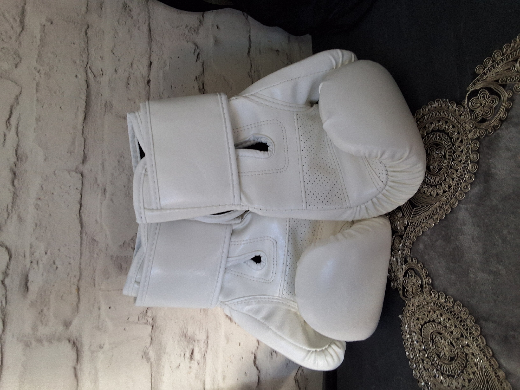A pair of white boxing gloves rests on a decorative, dark gray cloth with ornate gold trim. The gloves are positioned in the center of the frame, with the left glove slightly behind and to the left of the right glove. They appear to be made of a smooth material, possibly leather or faux leather, and have a quilted design on the palm area. The background is a textured, light-colored brick wall. The lighting suggests an indoor setting. No people are present in the image. The scene is static and appears to be a still life arrangement. The location is Unknown, Unknown.