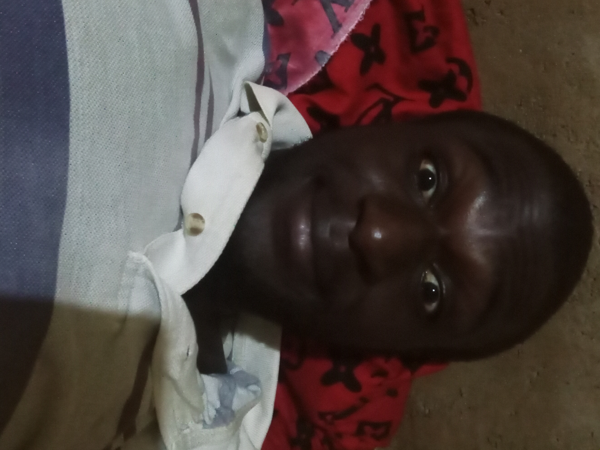 The image is a close-up, portrait-oriented photograph of a person lying down. The subject appears to be a young person with dark skin and dark hair. They are wearing a light-colored collared shirt with stripes in blue and dark red. The shirt is partially unbuttoned, revealing the collar and some of the chest area. The person is lying on a surface that appears to be a bed or a couch, with a red fabric featuring a black pattern of LV (Louis Vuitton) logos visible behind them.
The lighting in the image is dim, suggesting an indoor setting or possibly nighttime. The subject's gaze is directed slightly upwards and to the right of the camera, with one eye clearly visible and reflecting light. Their expression is neutral or perhaps slightly curious. The background is a rough, sandy-colored surface, possibly a floor or wall.
There is no discernible text in the image. The overall mood is intimate and candid, as if the photograph was taken spontaneously. The focus is on the subject's face and upper body, with the surrounding environment serving as a backdrop.