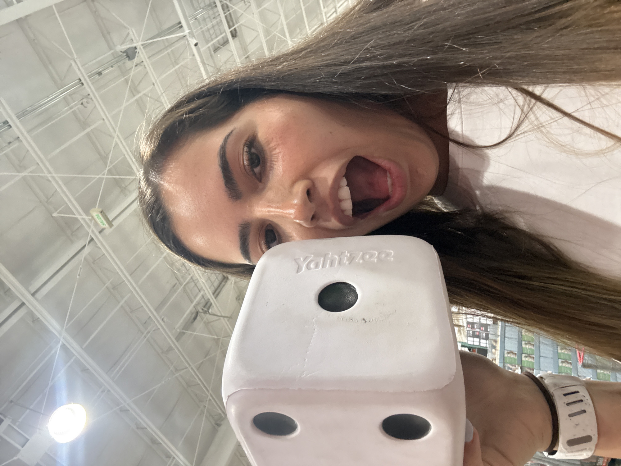 A woman holds a large, white Yahtzee die close to the camera, with her face visible above it. She has brown hair and her mouth is open wide. She is wearing a white shirt. The background shows a high, white ceiling with visible structural supports. A wristwatch is visible on her wrist. The location appears to be an indoor commercial space, possibly a store. The precise location in Puebla, Mexico cannot be determined based on the given image.