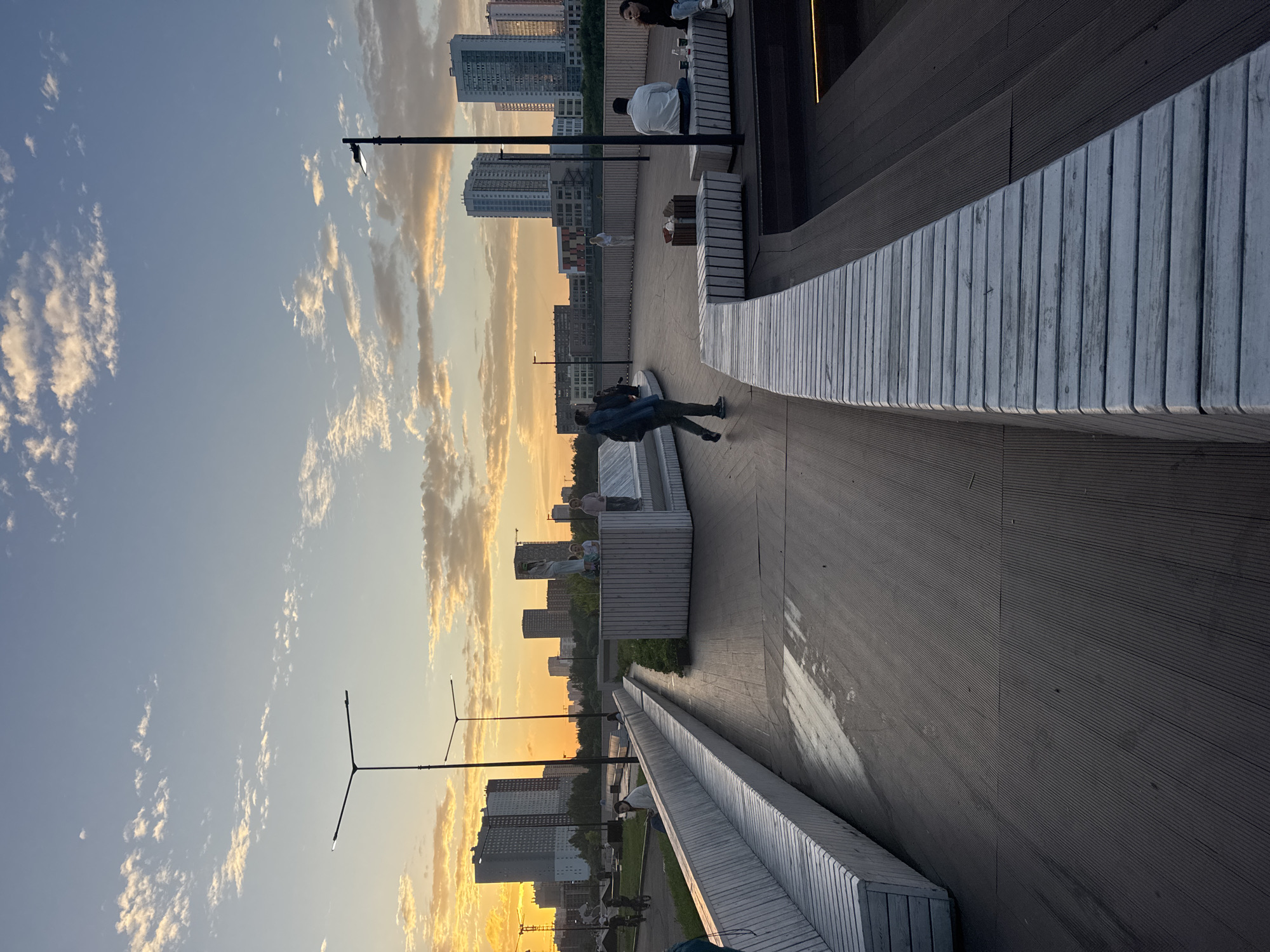 A person is captured in mid-air, appearing to jump or fall onto a wooden deck, dressed in a denim jacket and dark pants. The scene is an outdoor urban setting with modern, slatted wooden benches and walkways, illuminated by streetlights. In the background, a cityscape with tall apartment buildings and the sunset sky creates a backdrop of warm, orange and yellow hues mixed with light blue. The sky is dotted with wispy clouds, suggesting it's either late afternoon or early evening. Several other individuals are visible sitting on benches, seemingly enjoying the view or socializing. The overall atmosphere is calm and relaxed, bathed in the soft light of sunset. The location appears to be a contemporary park or promenade.