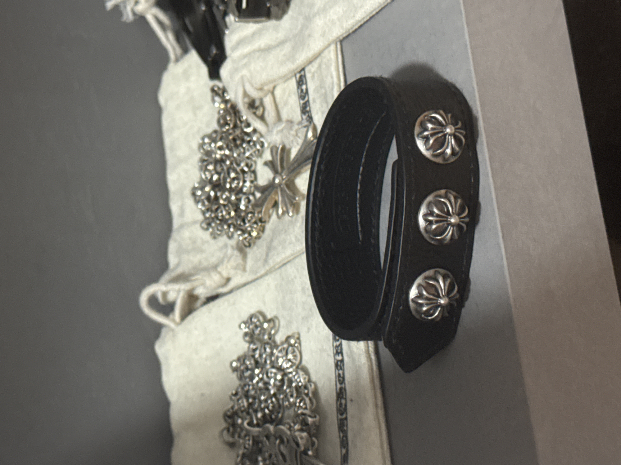 A close-up shot shows a black leather bracelet with three silver fleur-de-lis type emblems affixed to it. It is positioned next to a light-colored fabric pouch which appears to contain silver jewelry with similar fleur-de-lis designs, including necklaces and pendants. The background appears to be a combination of gray and dark shadow. There is no evidence to determine location in Burgas, Bulgaria.