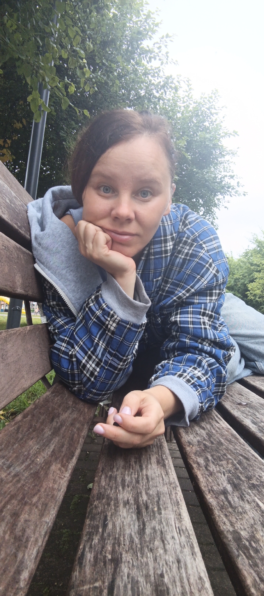 A medium shot shows a young woman lying on a wooden park bench in Chaikovsky, Russia. She is resting her head on her hand and looking directly at the camera with a neutral expression. Her hair is dark brown and pulled back. She is wearing a grey hoodie with the zipper partially open, revealing a blue and black plaid shirt underneath. The bench is made of weathered wood and is positioned outdoors, with trees and foliage visible in the background. The sky is overcast.