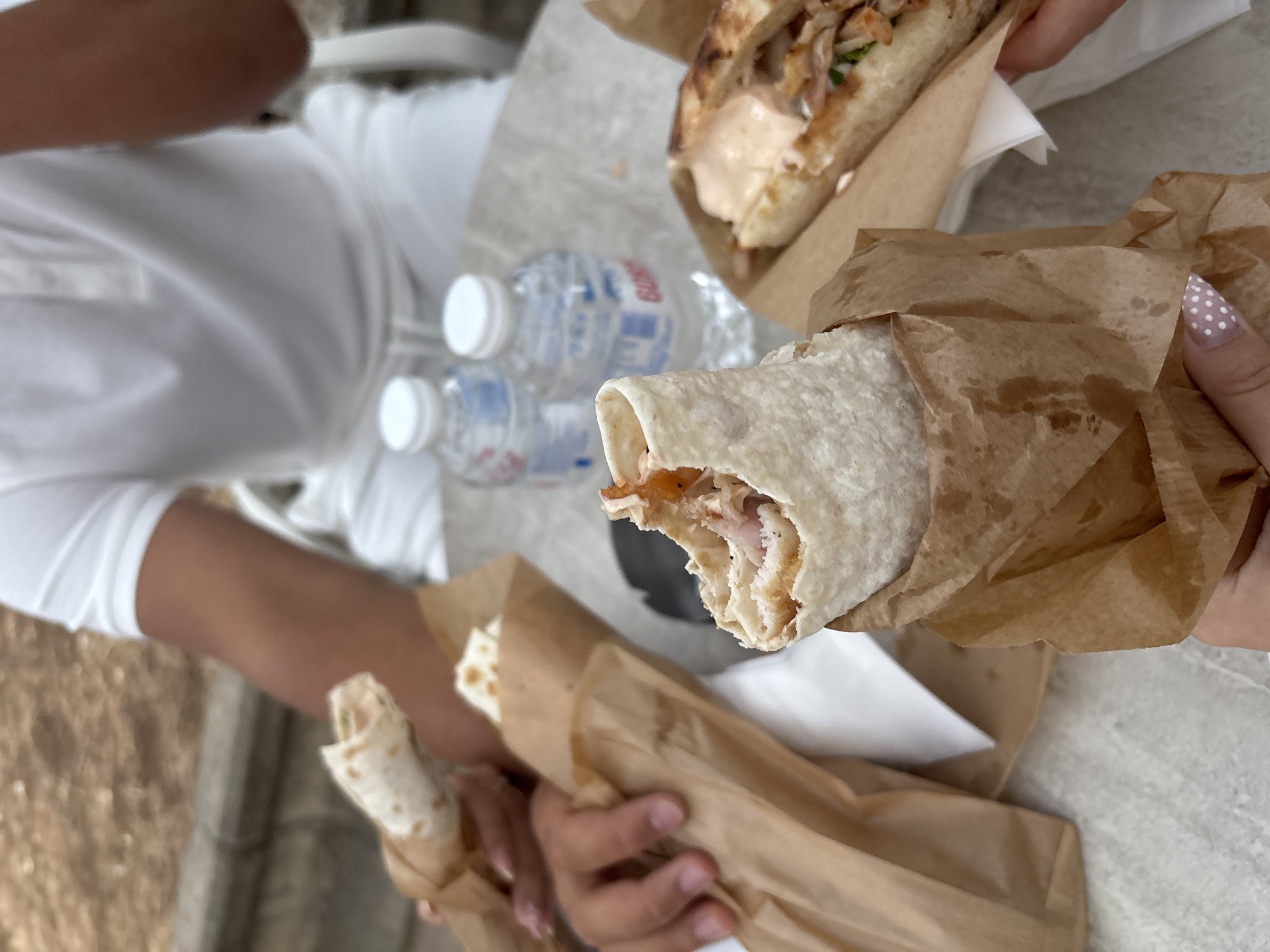 In Vratsa, Bulgaria, a close-up shot captures a casual meal outdoors. Several individuals, partially visible, are enjoying what appear to be wraps and sandwiches, each presented in brown paper packaging. Two plastic water bottles with white caps are visible on a textured surface, likely a table. The foreground is dominated by hands holding the food, one hand with a dotted nail design. The background shows hints of greenery and a blurred outdoor setting, suggesting a park or a patio. The overall impression is one of simple enjoyment of street food in a relaxed environment.
