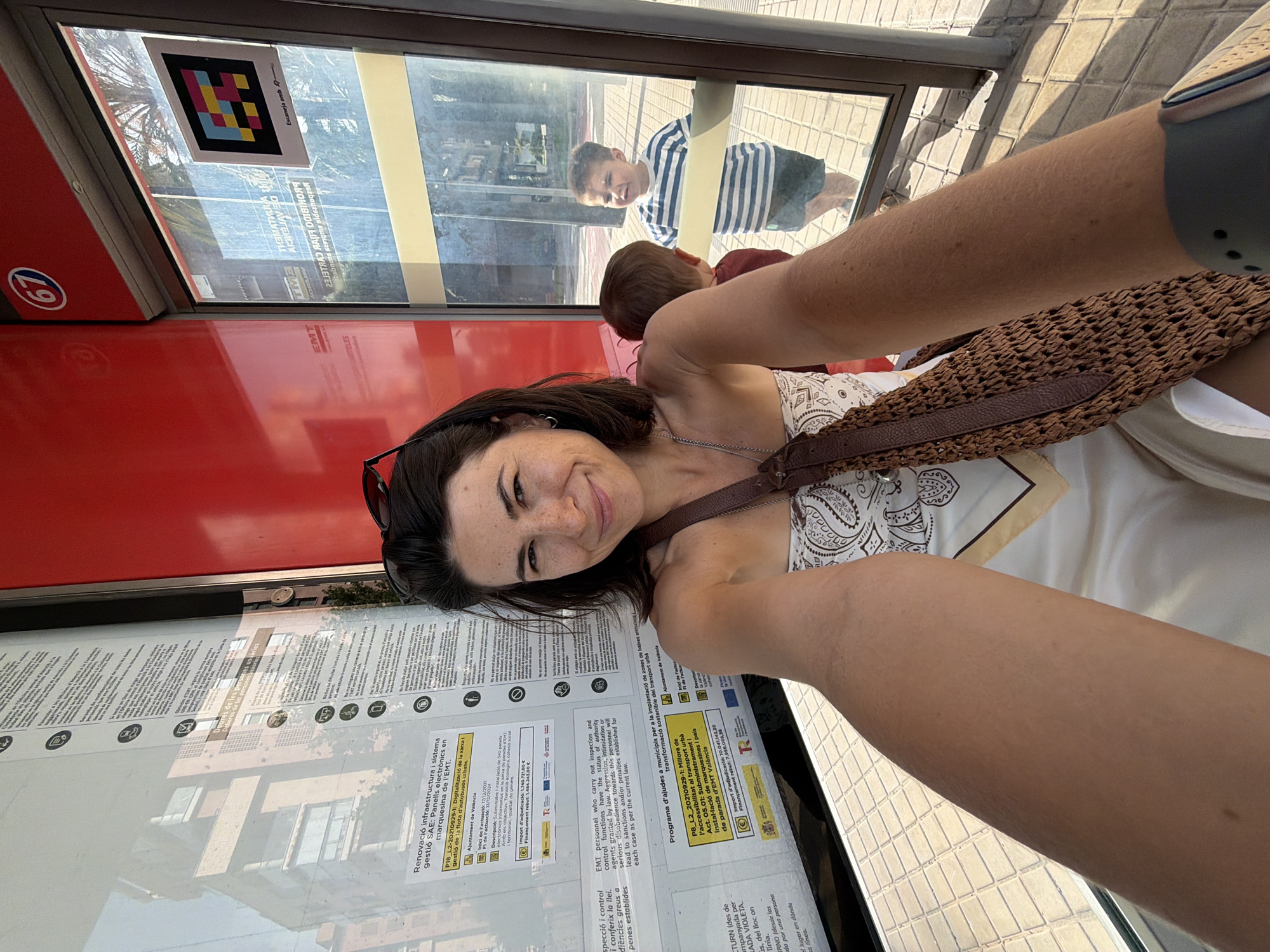 A woman is taking a selfie at a bus stop. She is wearing a strapless top and a patterned scarf. She has dark hair and is smiling at the camera. A young boy is visible in the reflection of the glass, wearing a striped shirt. The bus stop has a red exterior and a glass enclosure with informational posters. The ground is paved with tiles.