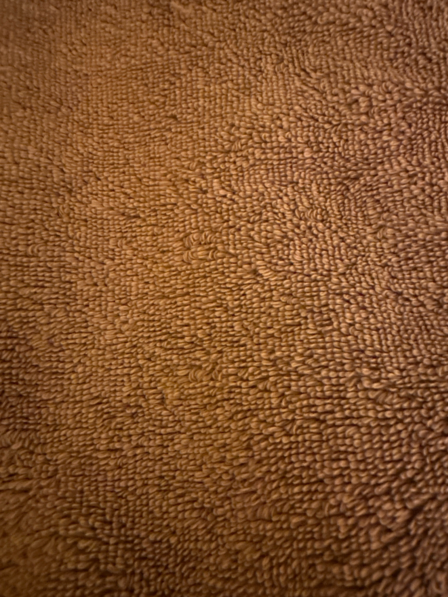 The image is a close-up, top-down view of a brown terry cloth towel. The texture of the towel is clearly visible, with individual loops of yarn forming a dense, slightly fuzzy surface. The lighting creates subtle shadows and highlights, emphasizing the texture and giving the towel a warm, earthy appearance. There are no people, objects, or environmental features visible that would suggest a specific location in Kathmandu Metropolitan City, Nepal, or any other location. The image focuses solely on the material and its texture.