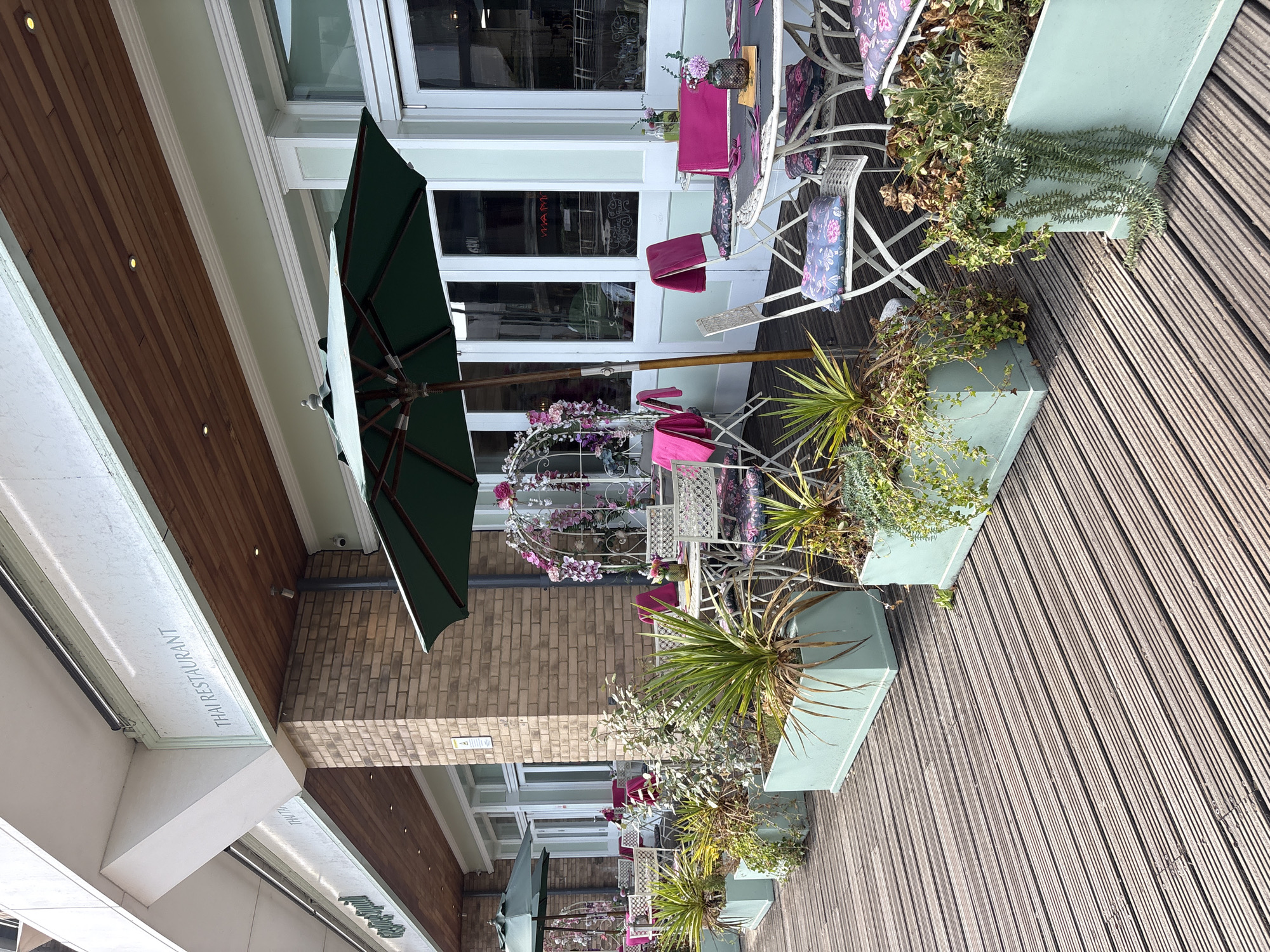 The image depicts the outdoor seating area of a Thai restaurant named "Thai Restaurant" located in Chelmsford, United Kingdom. The restaurant features a light green facade with large glass doors and windows. The outdoor seating area is furnished with white wrought-iron tables and chairs, adorned with floral-patterned cushions and bright pink napkins. Several planters filled with green and brown foliage are strategically placed along the wooden deck. Two green umbrellas provide shade over some of the tables. A decorative floral archway stands near the tables, adding a touch of elegance to the setting. The overall scene suggests a well-maintained and inviting outdoor dining space.