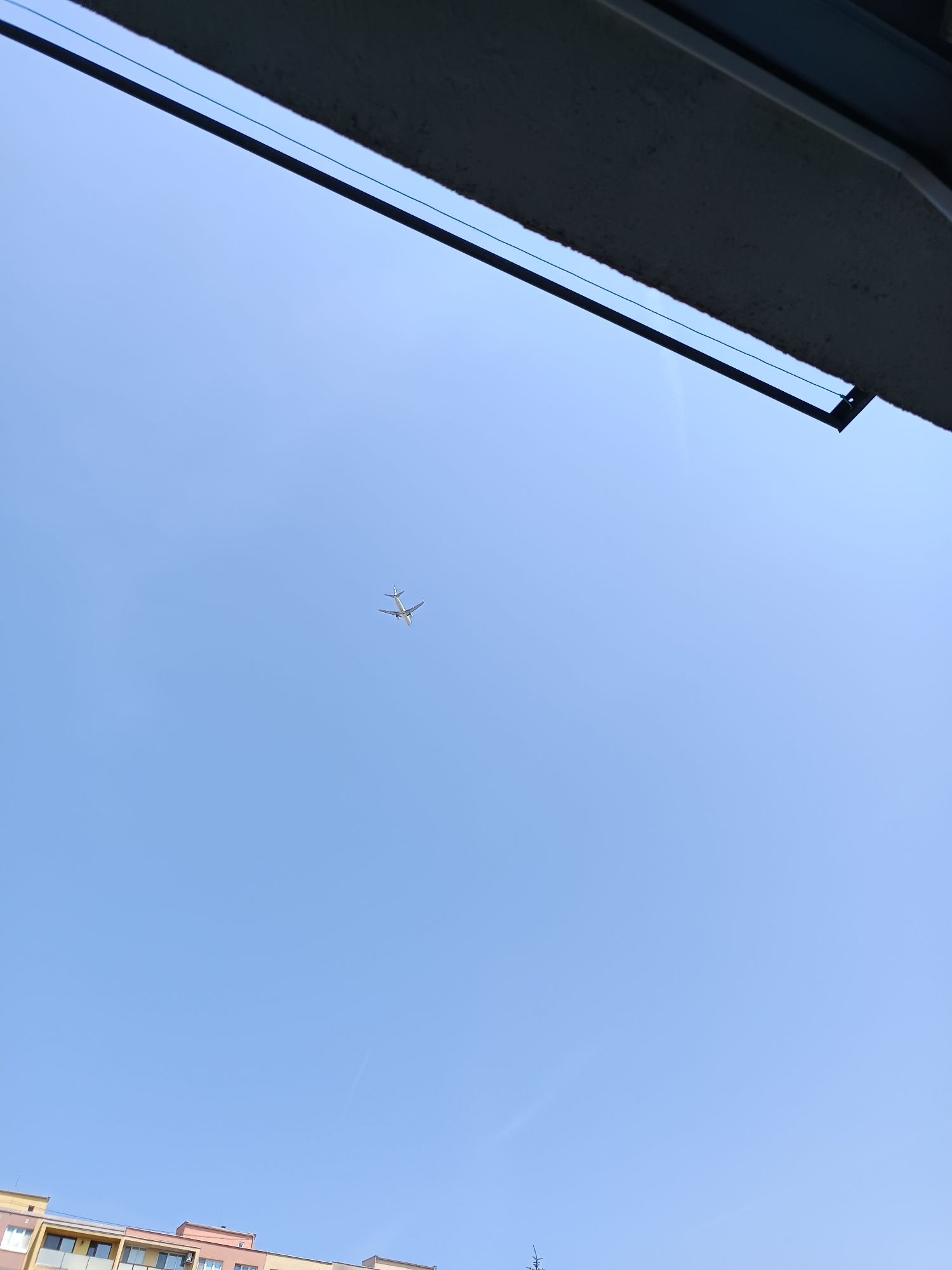 The image shows a clear, blue sky with a single airplane in flight. The airplane is white and appears to be a passenger jet, seen from below and slightly to the side. A portion of a building with multiple floors and balconies is visible at the bottom of the frame, suggesting an urban setting. The top of the image features the underside of an overhang or balcony, casting a shadow. The scene is likely from the city of Košice, Slovakia, given the presence of characteristic apartment buildings and the overall environment.