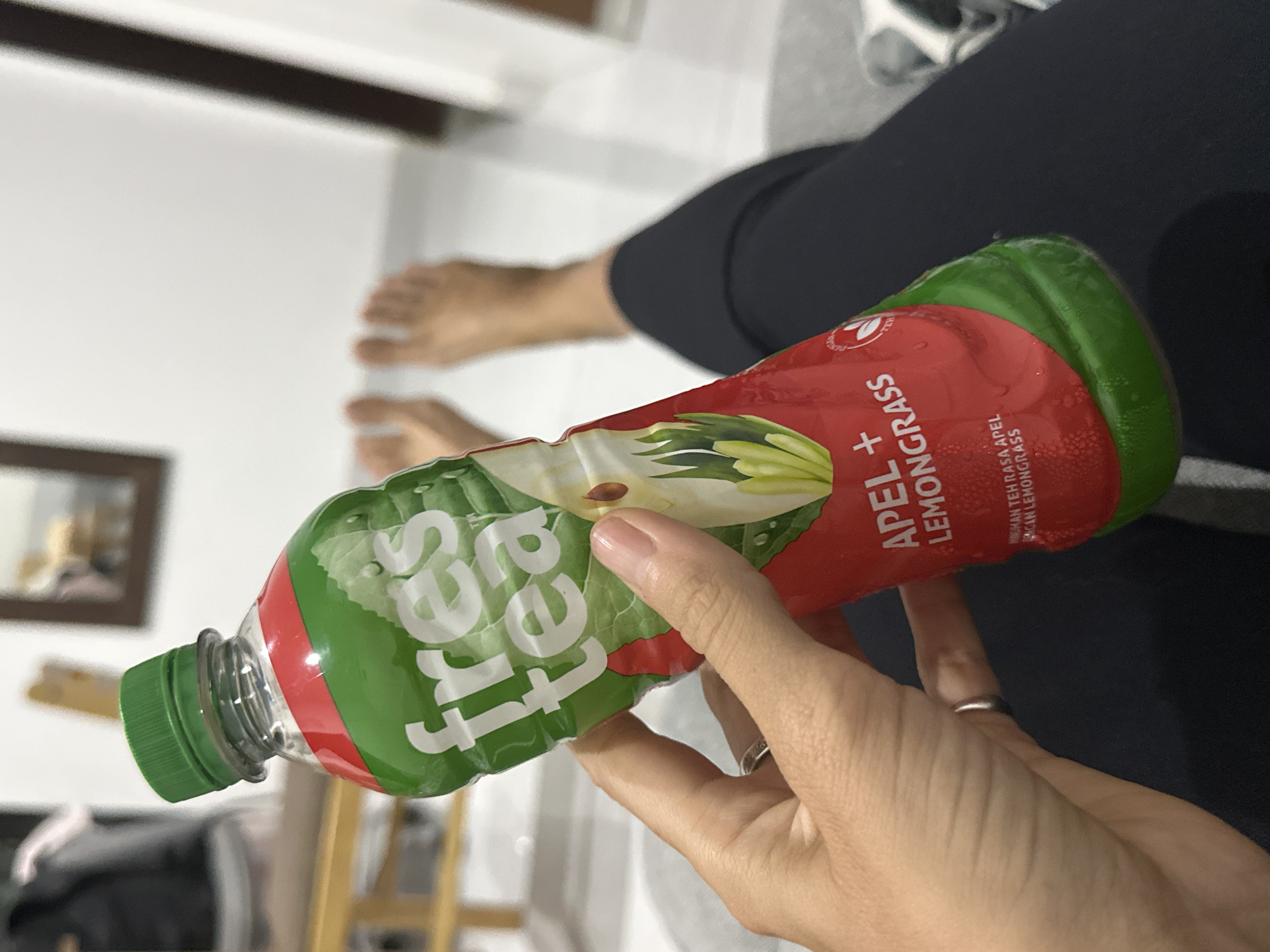 The image, taken indoors in Kuta Selatan, Indonesia, shows a person holding a bottle of "Fres Tea Apel + Lemongrass." The perspective is angled, with the bottle prominently featured in the foreground, held by the person's hand. The person's bare feet are visible in the blurred background, suggesting they are seated in a relaxed posture. They are wearing dark-colored pants. A silver ring is visible on one of their fingers.
The clear plastic bottle has a vibrant red and green label. "Fres Tea" is written in large white letters on the green section of the label, which also features illustrations of green leaves. The red part of the label states "APEL + LEMONGRASS" in white, accompanied by graphics of sliced apples and lemongrass stalks. Smaller Indonesian text, "MINUMAN TEH RASA APEL DAN LEMONGRASS" (Apple and Lemongrass Flavored Tea), is also visible on the label. The bottle has a green screw-cap, and condensation beads indicate the drink is cold.
The background is softly blurred, showing light-colored walls or flooring. A dark wooden frame, possibly a mirror or picture, is partially visible on the left. The indoor setting appears casual and well-lit. The activity depicted is simply holding the beverage, suggesting a moment of refreshment or rest.