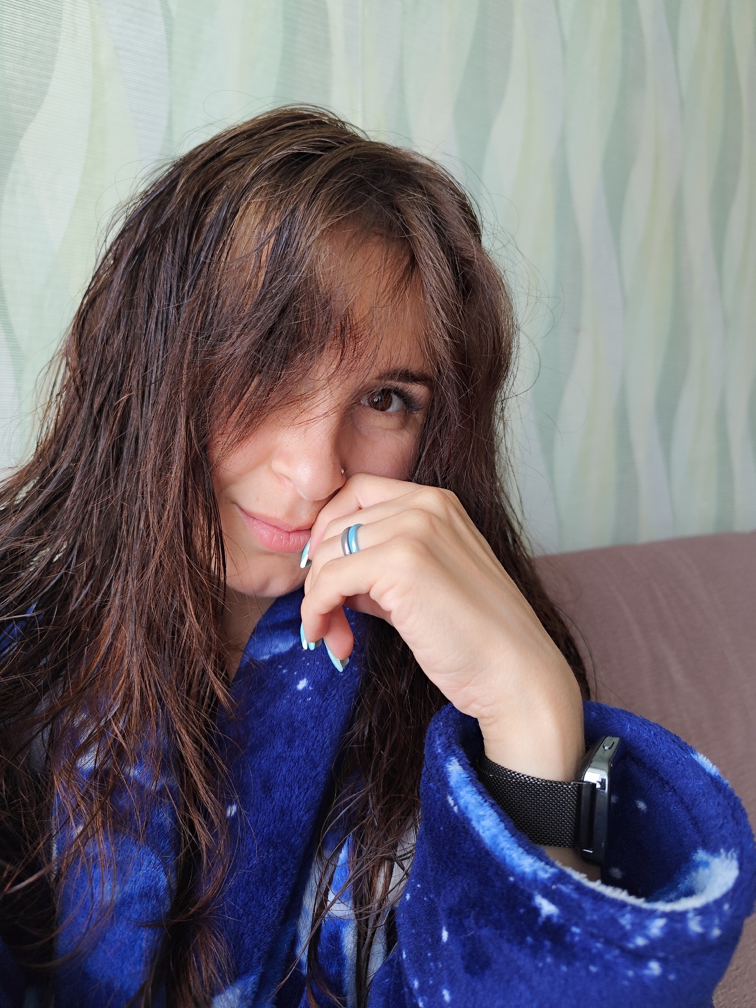 A woman with wet, dark brown hair partially obscuring her face is seen indoors. Her right eye is visible, and she has a nose piercing. She's resting her right hand near her face, wearing two rings on her ring finger. Her fingernails are painted light blue. She is wearing a blue, star-patterned robe. Her left wrist, partially obscured by the robe, reveals a black mesh watch band. The background consists of a wall with a light green wave-like pattern and a brown couch. There is no indication in the image that it was taken in Фоки, Russia.