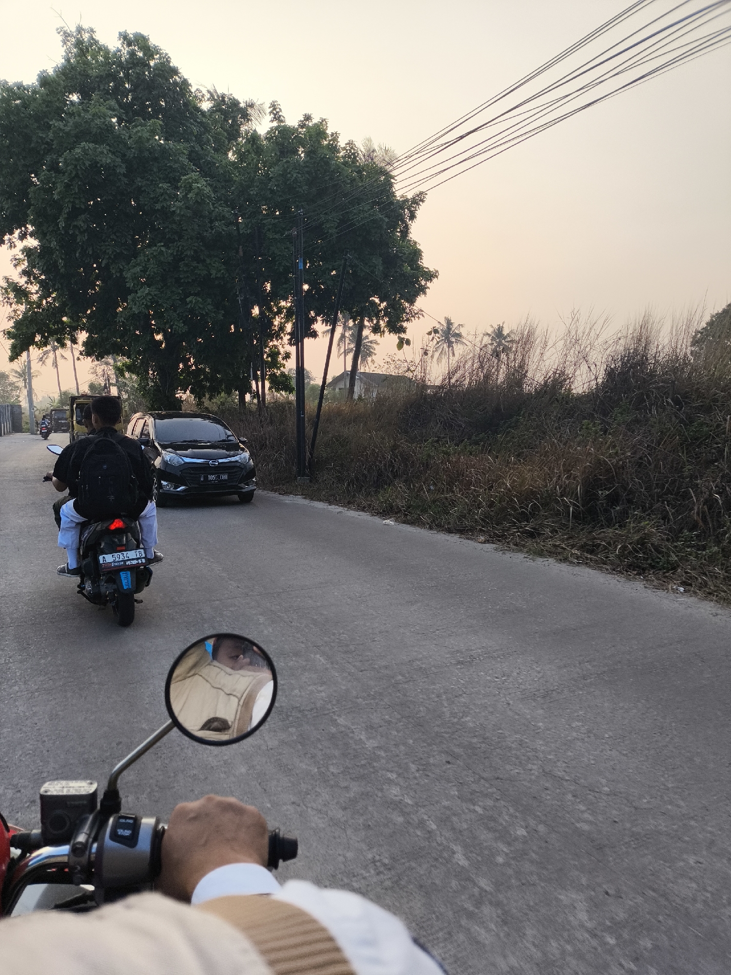 The image depicts a street scene in Cilegon, Indonesia, at dusk. In the foreground, a person's hand is visible, holding the handlebars of a motorcycle. A rearview mirror attached to the handlebars reflects a person wearing a light-colored vest. Following behind is another motorcycle, carrying two individuals. The rider in front has a backpack, and the passenger behind wears a light-colored shirt and white pants. A black car is in front of the motorcycles, and further down the road, a truck and another motorcycle are also visible. The environment is characterized by a paved road, tall trees, dry grass, and utility poles with overhead wires. The sky is a soft, warm gradient, suggesting either sunrise or sunset.