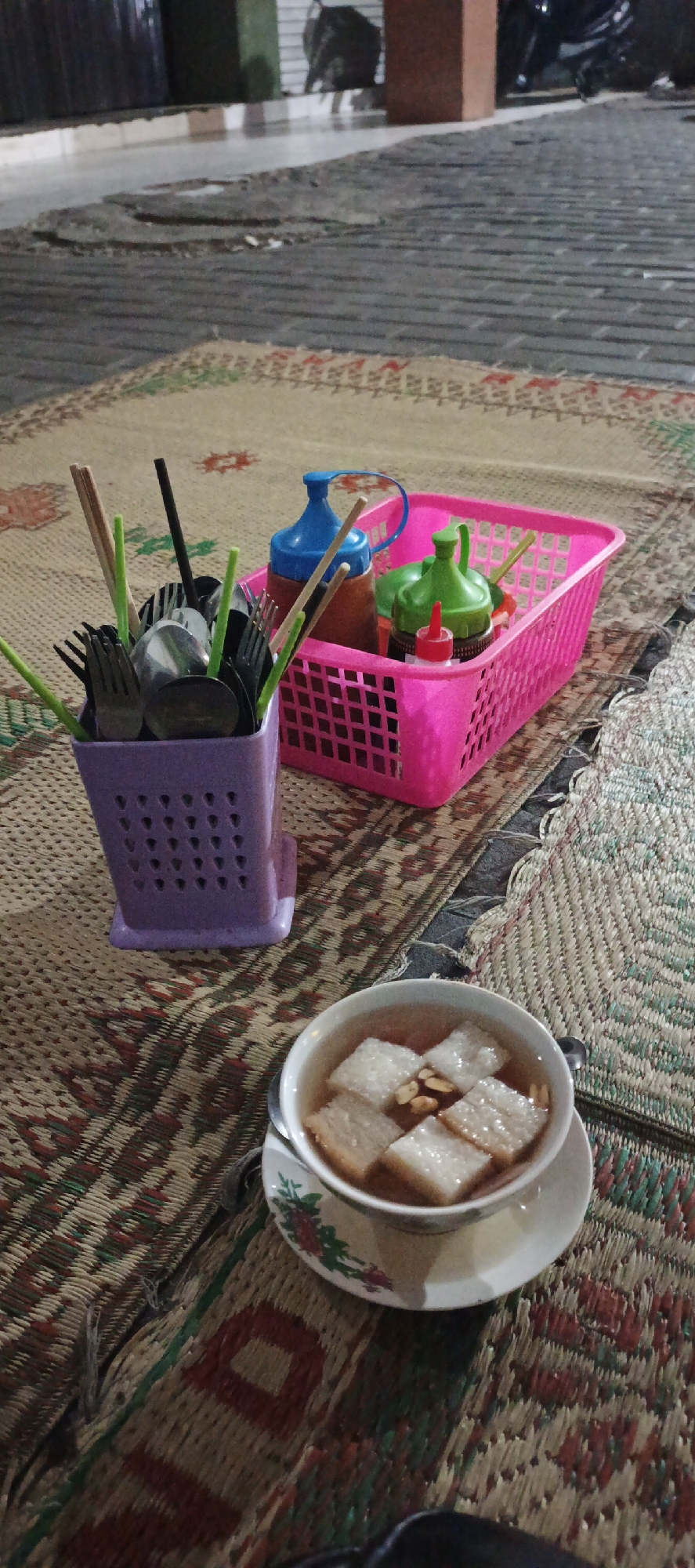 The image depicts a still life arrangement on a woven mat, likely set up for a meal outdoors. In the foreground, a white ceramic bowl contains a light brown broth with several rectangular pieces of white food and a few small, dark brown pieces that appear to be nuts or seeds. A spoon rests on the saucer beneath the bowl. To the left, a lavender-colored plastic utensil holder is filled with various cutlery, including forks, spoons, and chopsticks, as well as several green plastic straws or stirrers. Behind the utensil holder, a bright pink plastic basket holds an assortment of condiment bottles: one with a blue cap and a brown body, another with a green cap and a green body, and a small red-capped bottle. The background shows a textured ground that appears to be pavement or cobblestones, with elements of a building or structure and what looks like a motorcycle parked in the distance. The overall scene suggests a casual dining setting, possibly in a street food stall or an outdoor eating area in Godean, Indonesia, given the context.