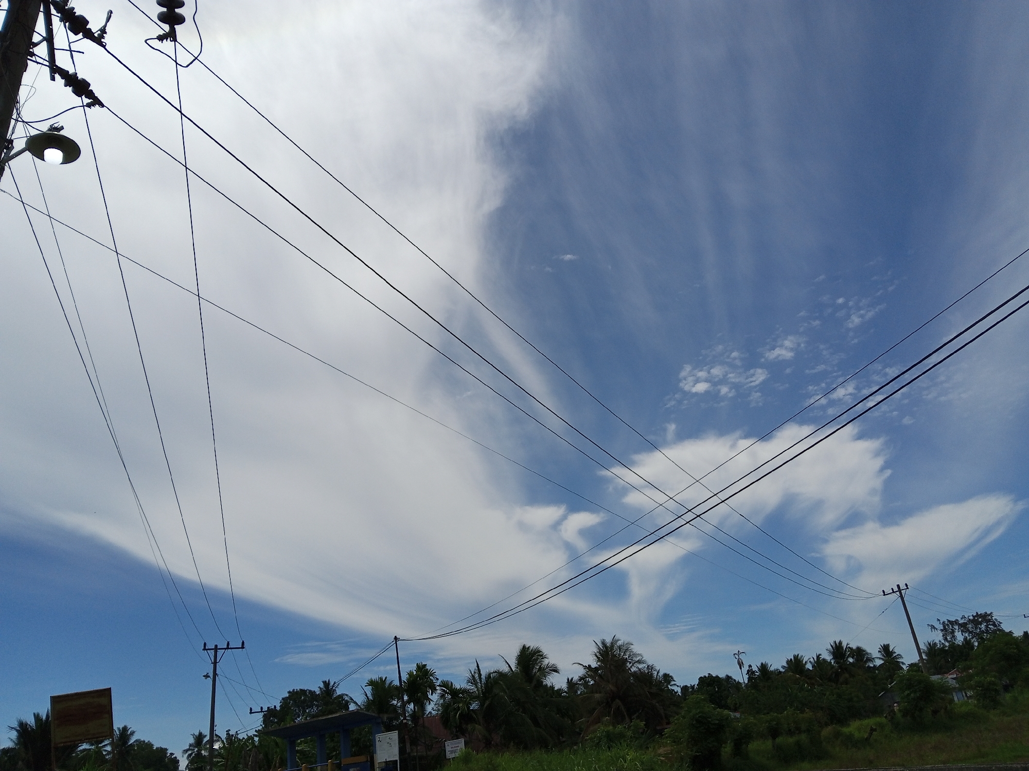 A low-angle shot captures a bright, partly cloudy sky over a dense green treeline in Cotjrat, Indonesia. Power lines crisscross the frame, suspended from utility poles. A street lamp is visible on the left, its bulb illuminated. The environment appears rural or suburban, with lush vegetation and tropical trees dominating the lower portion of the image. The sky transitions from a clear blue to areas filled with wispy white clouds, creating a dynamic visual contrast.