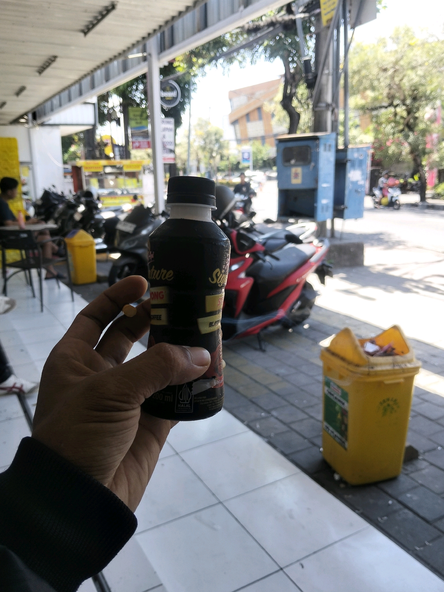 In Kuta, Indonesia, a person's hand holds a bottle of "Good Day Coffee Signature" beverage while a small, yellow pill rests on their index finger. The background reveals a street scene with parked motorcycles, a blue electrical box, and a yellow trash bin overflowing with debris. Sunlight filters through trees, casting shadows on the tiled ground. The overall setting appears to be an outdoor seating area of a cafe or food stall.