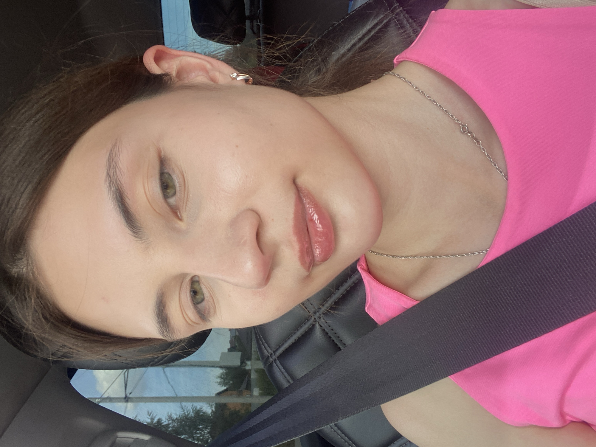 A young woman with light skin, light brown hair, and green eyes is in the passenger seat of a car, looking at the camera. She is wearing a bright pink tank top and a silver chain necklace. Her seatbelt is across her body. The background shows a window with trees and a blue sky with clouds.