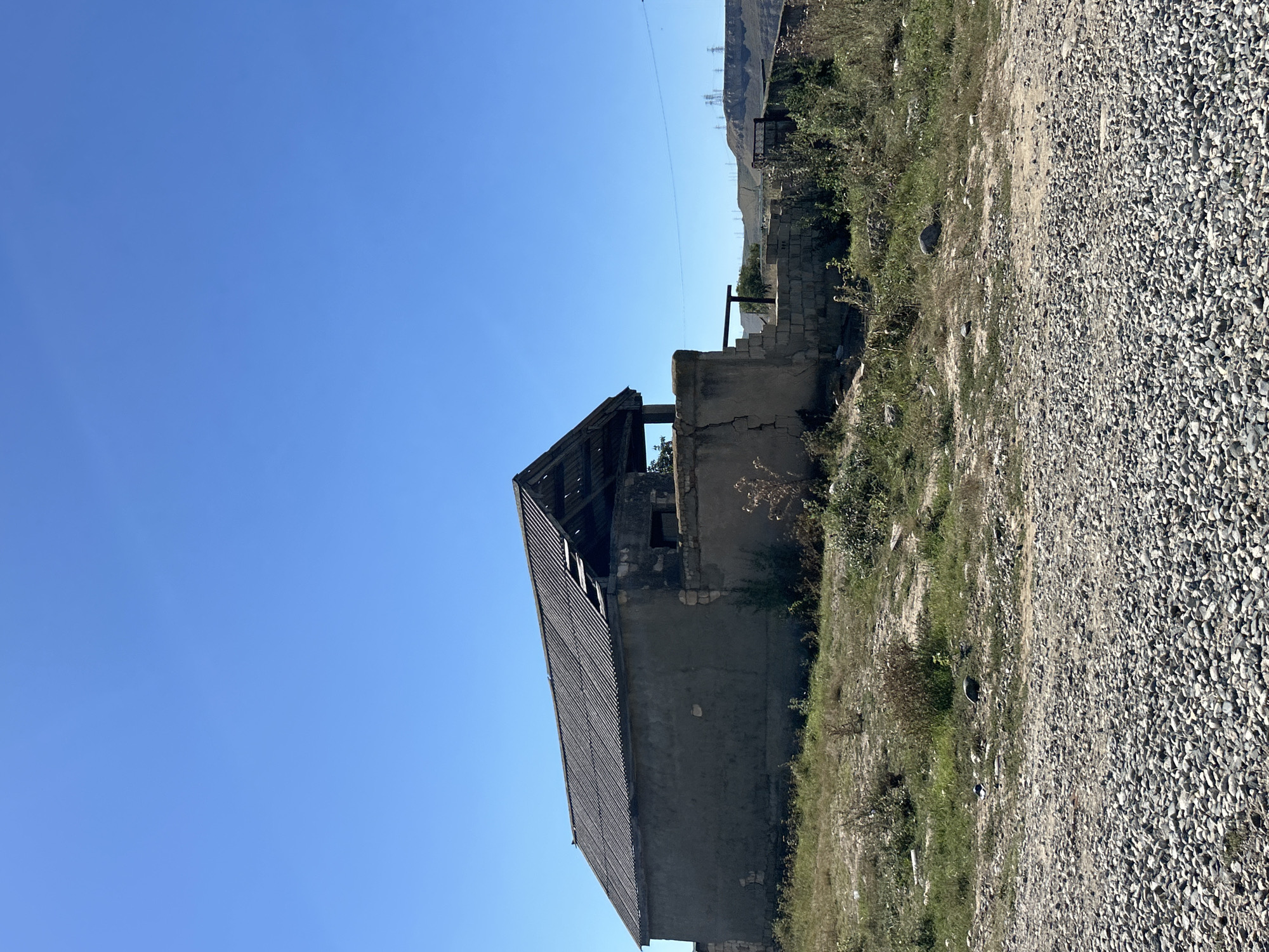 In an outdoor scene under a clear blue sky, a dilapidated structure stands on a patch of dry, overgrown grass and gravel. The building, seemingly abandoned, has a corrugated roof with sections missing, exposing the wooden framework beneath. The walls are rough, unpainted concrete and stone. In the middle ground, there are low-lying shrubs and dried vegetation. The foreground consists of a gravelly surface. In the far background, hills are visible, along with power lines and transmission towers, suggesting a rural or semi-rural setting on the outskirts of a settlement. The overall impression is one of neglect and disrepair. There are no people or animals visible in the frame.