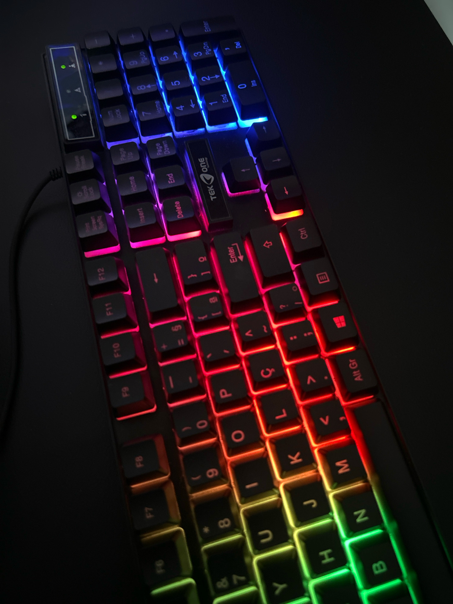 An overhead, close-up, low-angle shot shows a black gaming keyboard with RGB lighting on a dark surface. The keyboard is angled with the top-left corner closest to the viewer. The keys are illuminated with a gradient of colors, from blue on the top row, to pink and red in the middle rows, and transitioning to yellow, green, and a light blue on the bottom rows. Some of the keys are visible, including F1 through F12, the number pad keys, and alphanumeric keys. A cable connects to the left side of the keyboard. The lighting creates a dramatic effect, highlighting the contours of the keys. The image lacks any visual cues to suggest a specific location within Teixeira de Freitas, Brazil.