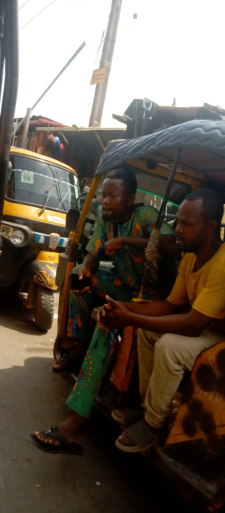 The image depicts a street scene in Onitsha, Nigeria, likely during the daytime under bright, possibly hazy or overcast sky conditions. Two men are seated in a yellow auto-rickshaw, commonly known as a "keke Napep," appearing to be waiting or resting.
The man positioned more prominently on the left wears a vibrant green long-sleeved top and matching trousers with an orange and black abstract pattern. He has a dark beard and mustache and holds a black mobile phone in his left hand. His left foot, clad in a black flip-flop, rests on the ground.
To his right, the second man wears a plain yellow short-sleeved t-shirt and light-colored trousers. His hands are clasped in front of him, and he wears dark slides or sandals. He also has a dark beard and is looking towards the right side of the frame. Both men appear calm and relaxed.
The yellow keke Napep shows signs of wear, particularly on its front section. The brand "PIAGGIO" is visible on its front, above a blue and white striped design and a circular emblem. The vehicle's single front tire is partially visible.
In the background, behind the keke, blurred structures suggest buildings or market stalls, indicating a bustling urban or commercial setting. A tall street pole extends upwards on the right side of the frame. Attached to this pole is a rectangular yellow sign with black text, which reads "NO WAITING KEEP MOVING," signifying a traffic or parking regulation. Overhead wires are also visible against the bright sky. The ground beneath the keke is a dark, paved surface.