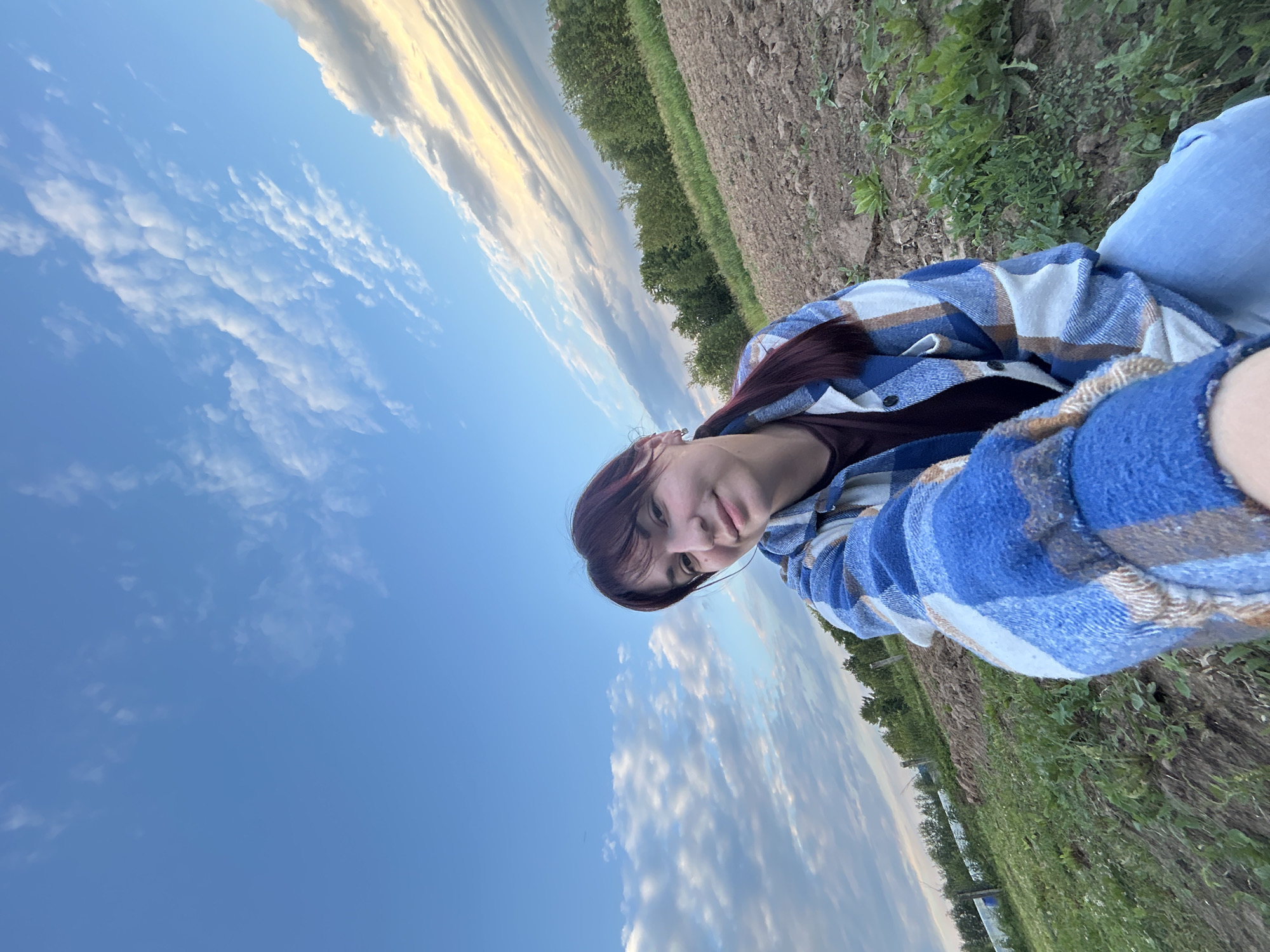 A young woman with dark hair pulled into a ponytail and bangs is pictured in a selfie taken outdoors in the city of Fokino, Russia. She is wearing a blue and white plaid shirt over a dark t-shirt and blue jeans. The background shows a field with cultivated soil, green grass, and a line of trees in the distance under a bright blue sky with scattered white clouds. The lighting suggests it is either early morning or late afternoon.