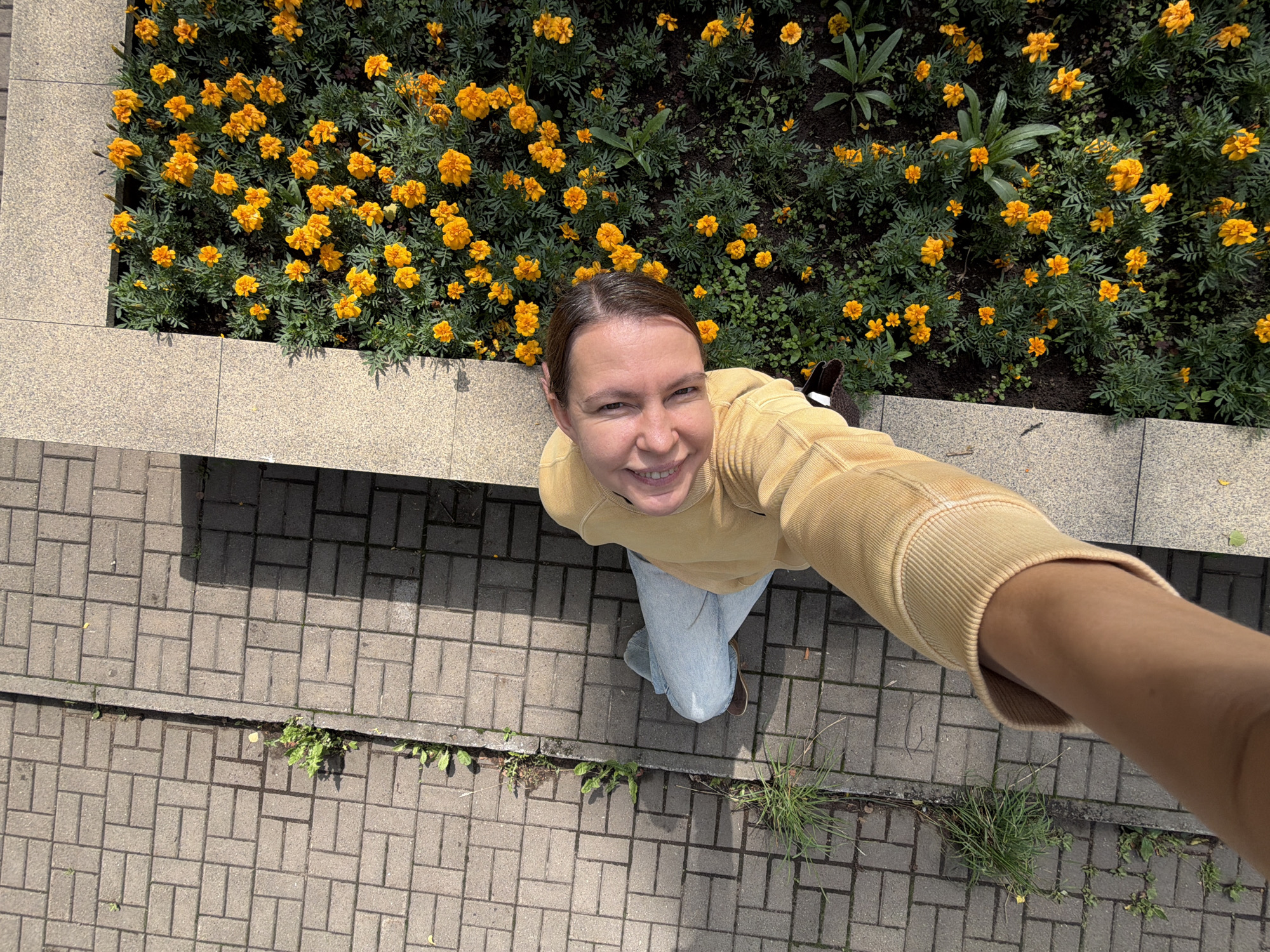 A young Caucasian woman with dark hair pulled back, wearing a yellow hooded sweatshirt and light blue jeans, is taking a selfie outdoors. She is looking up and smiling at the camera, with her left arm extended. The woman is positioned on a paved walkway made of grey rectangular bricks, which has a strip of grass growing in the cracks between the bricks. To her right is a raised flower bed filled with numerous bright orange marigolds and green foliage, bordered by beige stone tiles. The scene appears to be in an urban setting, possibly a park or public square, with sunlight casting shadows on the ground. The image was taken in Chaikovsky, Russia.