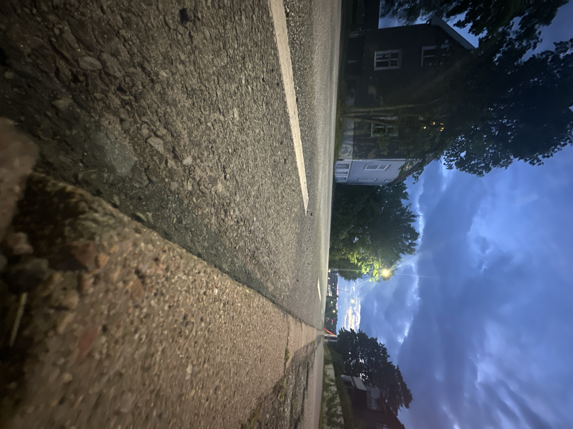 A low-angle, close-up shot captures a paved road at twilight, with a concrete curb in the foreground. A white line runs along the road, dividing the lanes. To the left, a two-story house with white-trimmed windows is visible, partially obscured by trees. The sky above is dark and cloudy, with a streetlamp casting a bright yellow glow. The scene is urban and deserted, with no people or vehicles present. The overall mood is serene and slightly ominous, due to the dramatic sky and low light conditions.