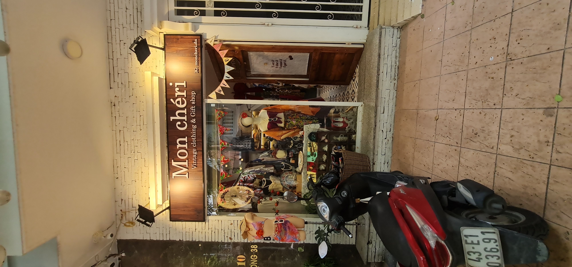 The image is a storefront of "Mon chéri," a vintage clothing and gift shop, likely taken in Đà Nẵng, Vietnam, based on the street number and vehicle license plate. The shop has a wooden-framed sign with the name "Mon chéri" in bold letters and "Vintage clothing & Gift shop" underneath. A small sign above the store reads "IG: moncheri.dn."
The shop window display is crowded with various items, including mannequins dressed in vintage clothing, hats, and accessories. There is a welcome banner hanging above the window display. The entrance is partially open, revealing the interior of the shop.
A parked motorcycle is in front of the shop, partially blocking the view of the storefront. The motorcycle is mostly black, with a red rear fender and a license plate visible: "43-E1 036.91." The motorcycle partially obscures the address on the building which reads, "10 ĐƯỜNG 38 B".
The storefront is white brick and has two spotlights that illuminate the shop's sign. The surrounding area appears to be tiled. The image is rotated 90 degrees clockwise.