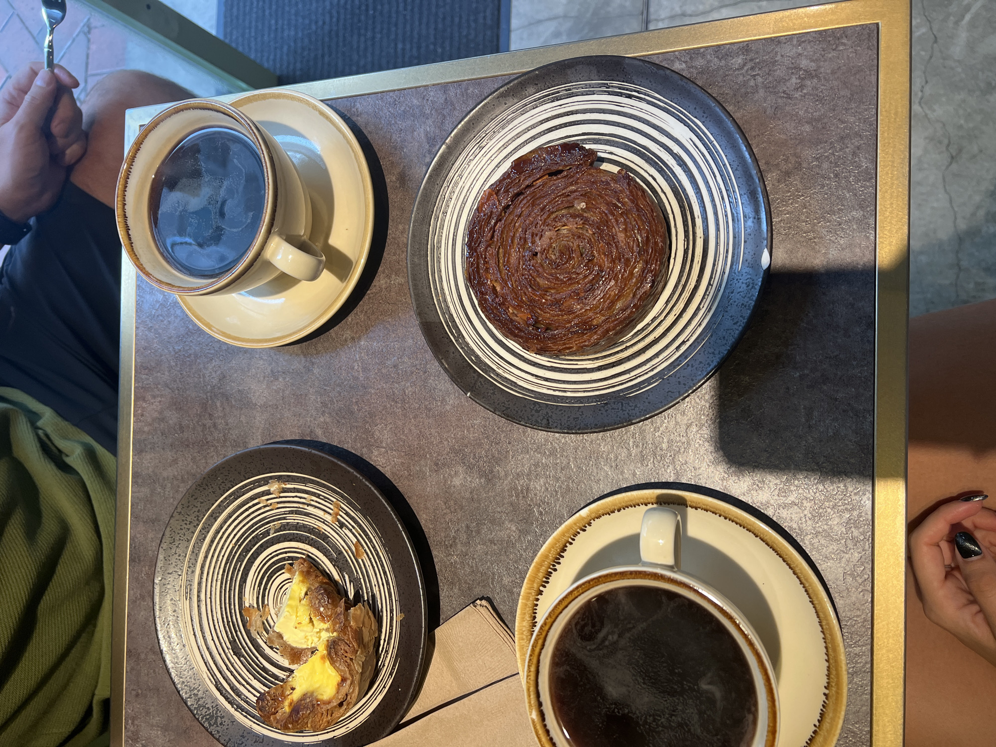 A top-down, medium shot captures a table setting for two, likely in a Hong Kong cafe. Two ceramic coffee cups, one lighter beige with a brown rim and the other slightly darker with a beige rim, sit on matching saucers filled with black coffee. The left cup is positioned on the bottom left of the frame, while the right cup is on the upper right. Each cup has a handle.
In front of the left coffee cup, on a dark, circular plate with a white concentric circle pattern, is a partially eaten pastry with flaky crust and a yellow custard filling, resembling an egg tart. A brown paper napkin is placed to the left of this plate.
To the right of the coffee cups, on another dark, circular plate with a similar white concentric circle pattern, sits a whole, circular pastry with a rich brown, layered, and swirled appearance, possibly a croissant or kouign-amann.
The table surface appears to be dark, possibly a speckled laminate or stone, with a thin gold trim along the edges. In the background, a person wearing a green top and dark shorts is partially visible, with their right hand holding a fork. Another person's hand with dark, glossy nail polish is visible in the bottom left corner of the frame. The environment suggests an indoor setting with natural light casting shadows across the table.