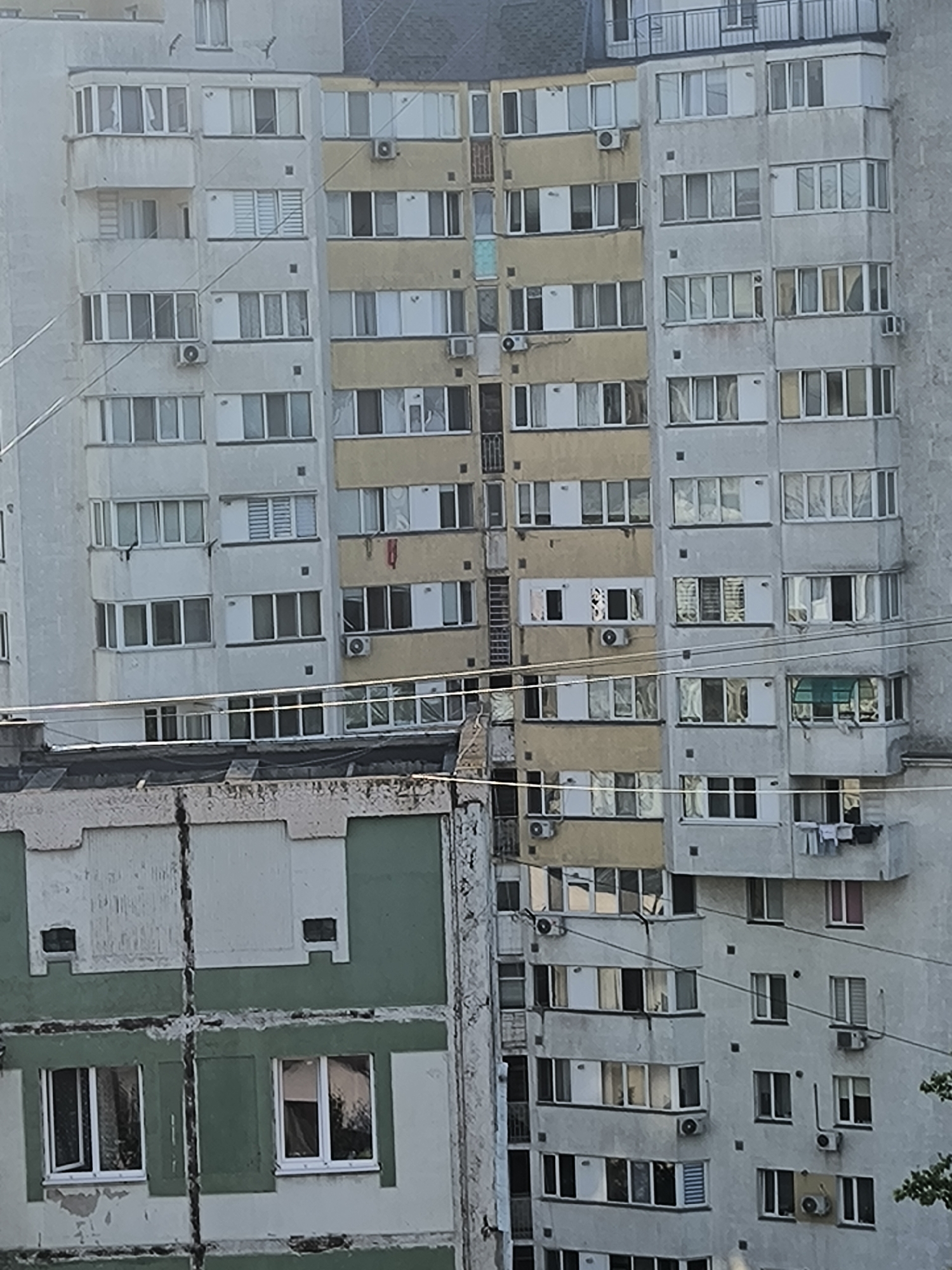 This is a medium-distance, eye-level photograph of a multi-story apartment building in Chișinău, Moldova. The building is constructed from concrete and appears to be of Soviet-era architecture. It has a light-colored, weathered facade with numerous windows, some of which are covered with blinds or curtains. There are balconies on several floors, some enclosed and others open. Air conditioning units are visible attached to the exterior walls. The building is densely packed with residential units, suggesting it is an apartment block. Some sections of the building are painted yellow, while others are a light gray. The lower portion of the image shows a different, less well-maintained building with a green and white facade, featuring two windows with white frames. The foreground is dominated by power lines strung across the frame, obscuring parts of the buildings. The sky is not visible, and there is no greenery in the immediate vicinity of the buildings. The scene appears to be an urban residential environment.