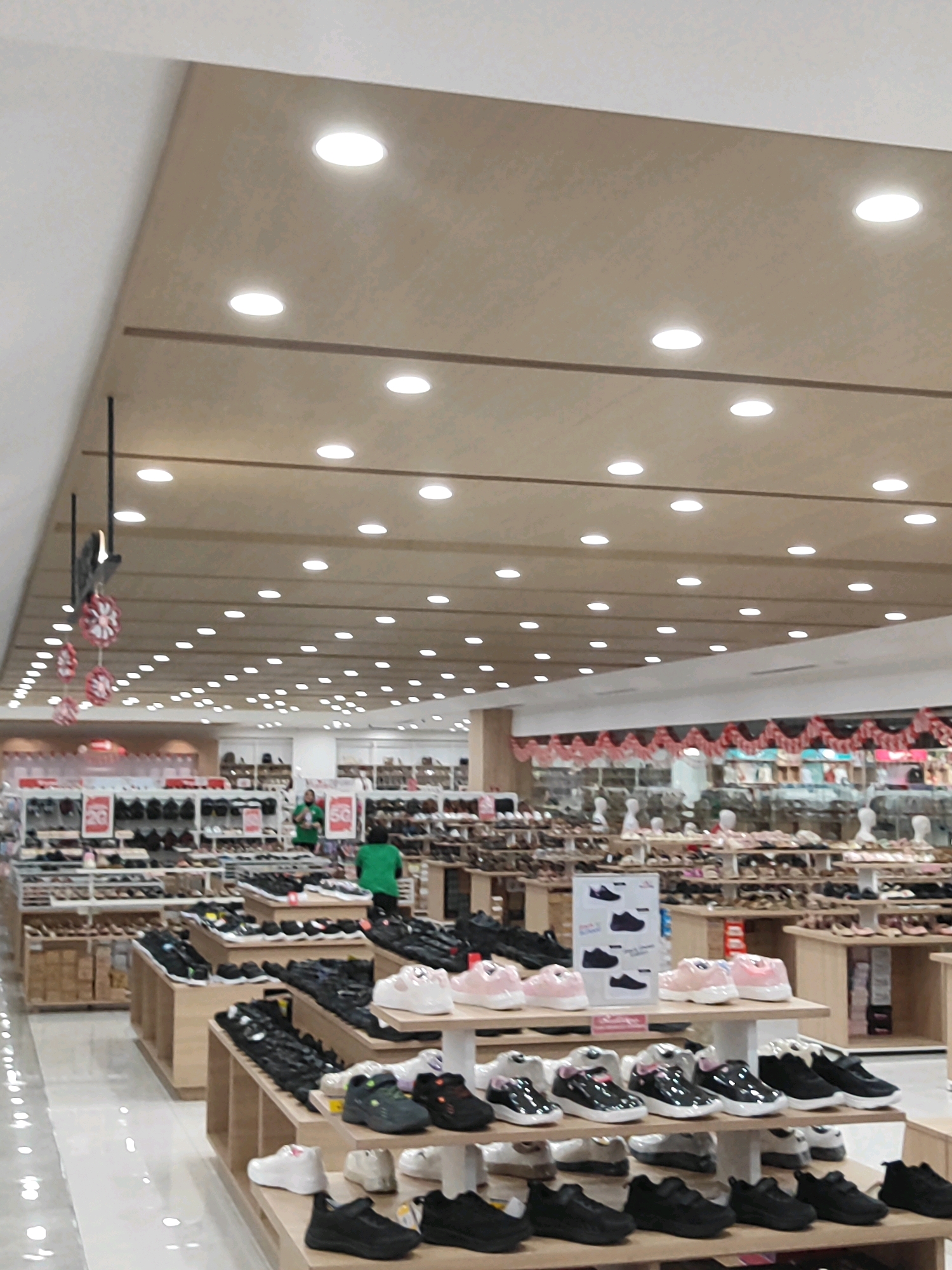 This image depicts the interior of a shoe store located in Sukowiryo, Indonesia. The store is brightly lit by numerous recessed ceiling lights. The foreground displays various shelves and displays of shoes, organized by type and color. There are multiple racks of footwear, including athletic shoes, casual shoes, and children's shoes. In the midground, there are two individuals, one wearing a green shirt and another in a darker green shirt, appearing to be employees or customers. They are standing near a display that has red and white signage with the number "50" visible, possibly indicating a sale. The background reveals more rows of shoe displays, suggesting a large retail space. Decorative red and white pennant banners are hung across the top of some of the displays. The flooring is a light-colored, possibly tiled or polished concrete surface. The overall impression is of a busy retail environment focused on footwear.