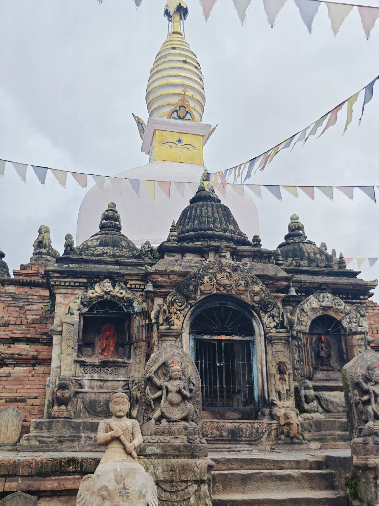 This is a medium shot, eye-level, outdoor photograph of the Chilancho Vihar in Kirtipur, Nepal. The stupa, topped with a golden spire and painted with Buddhist eyes, is partially visible against an overcast sky. Colorful prayer flags are strung across the frame, partially obscuring the stupa and the lower temple structures. The temple features intricate stone carvings and multiple statues, including a seated figure in the foreground, a dancing deity to the right, and several smaller figures adorning the facade. Brick walls are visible on the left side of the image. The overall atmosphere is solemn and spiritual.
