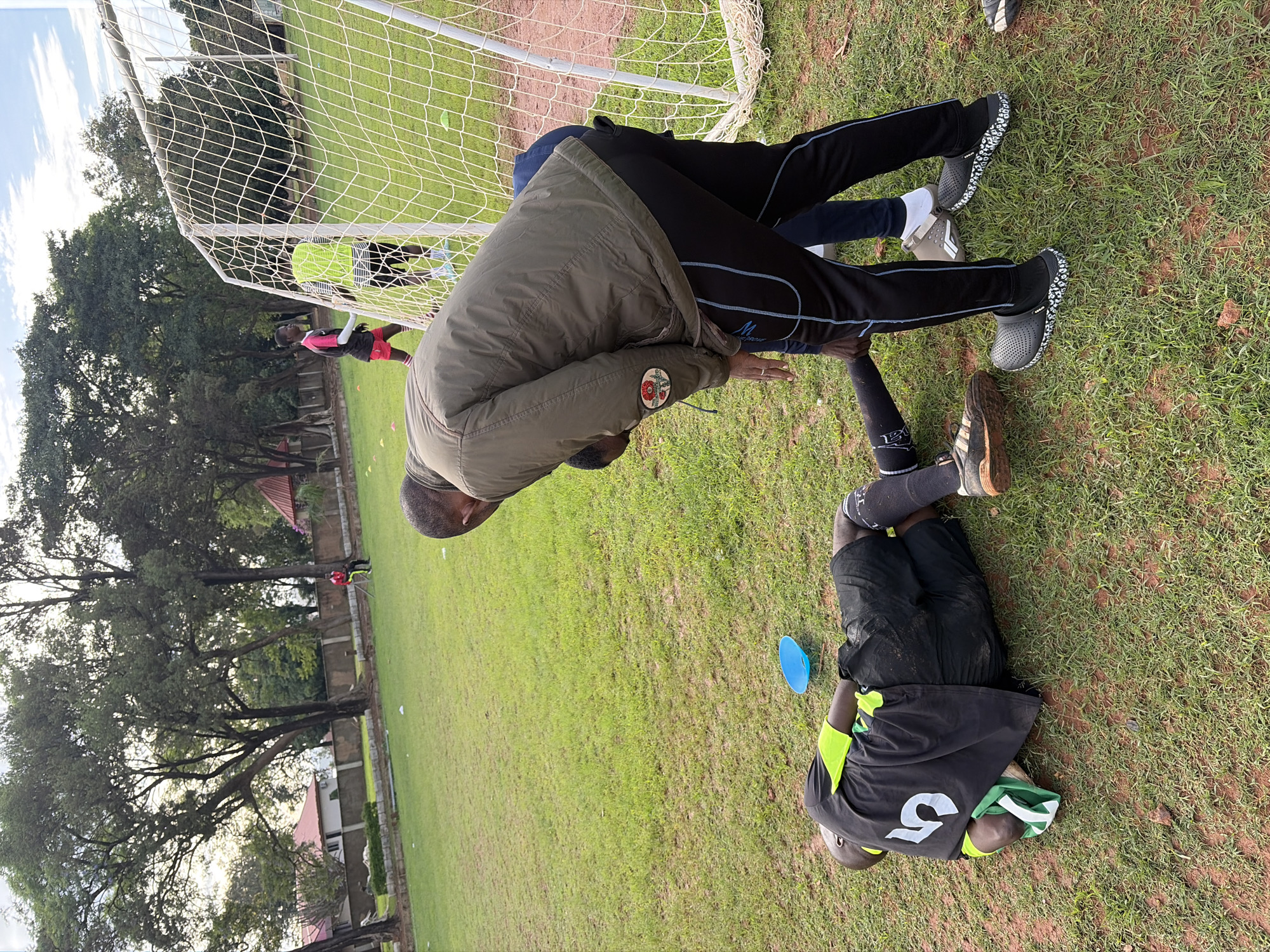 This image depicts an outdoor scene on a grassy field in Jos, Nigeria, likely a football pitch or training ground.
In the foreground, a man is lying on his left side on the grass, wearing a black football jersey with a large white number "5" on the back, black shorts, and black socks. His jersey features bright yellow-green sleeves, and a green and white striped cloth is partially visible near his head. He wears a dark-colored athletic shoe on his right foot. Adjacent to him, a light blue, inverted cone-shaped marker rests on the grass.
Standing over the man on the ground and bending forward is another man, dressed in a dark olive green jacket, black athletic track pants with light blue piping, and black patterned clogs worn over black socks. His right hand is positioned near the head or upper torso of the man on the ground. Partially visible behind him, another individual wears blue pants and white socks with grey and white athletic shoes, also leaning forward and looking down at the man on the ground.
In the mid-ground, to the right, a white metal football goal with a white net is positioned. Two individuals are near the goal: one wearing a black and pink jersey with red shorts stands to the left of the goal frame, while another wearing a bright yellow-green sleeveless bib over a striped shirt is positioned inside the goal area. Several small orange cone markers are scattered across the green grass further back from the foreground figures.
The background consists of numerous tall, leafy green trees forming a dense line. Beyond the trees, several buildings with reddish-brown roofs and light-colored walls are visible, suggesting a residential or institutional area. The sky above is light, with scattered white clouds, indicating daylight conditions. The ground primarily consists of green grass interspersed with patches of reddish-brown soil, particularly in the immediate foreground and near the goal area.