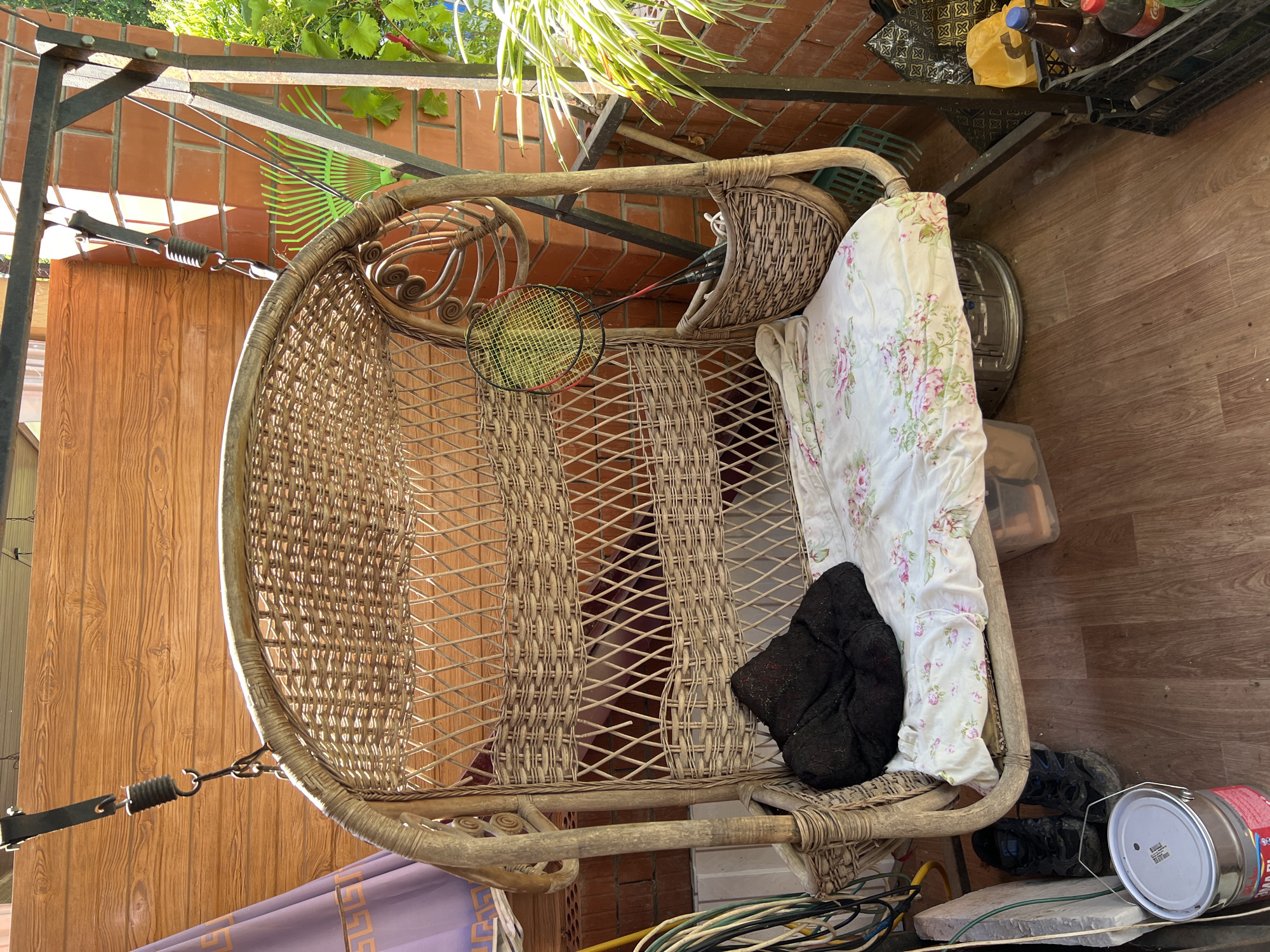 This image depicts a wicker swing chair situated on a wooden porch or balcony. The chair is equipped with a floral-patterned cushion and a black cushion. Hanging from the chair's frame are two badminton rackets and a shuttlecock. To the left of the swing, there is a coiled green hose and a metal bucket with a red label. Underneath the swing, a pair of muddy black shoes are visible. The background consists of a wooden wall and a brick wall, with some greenery and gardening tools, including a rake, also present. The scene appears to be an outdoor area of a private residence. The specific location in Goryachiy Klyuch, Russia, is not explicitly identifiable from the image content itself, but the context suggests a warm climate given the outdoor setting and foliage.