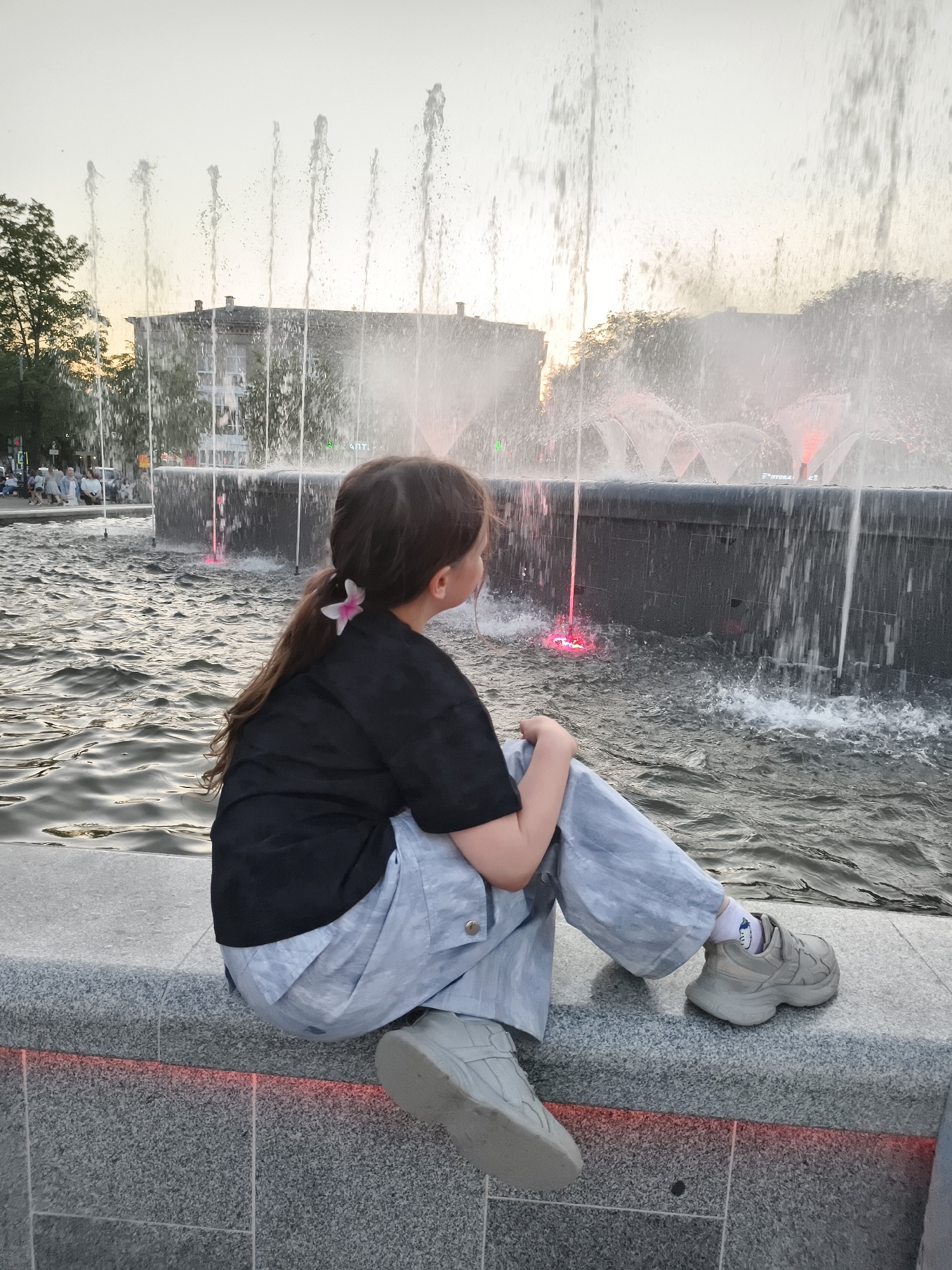 A young girl with brown hair pulled back, adorned with a small white flower, sits on a gray stone ledge, facing away from the viewer and toward a large, ornate fountain. She wears a black short-sleeved shirt and light blue, wide-legged pants, with chunky gray sneakers and patterned socks visible on her feet. The fountain is in the background, featuring numerous water jets spraying high into the air, with some illuminated by red lights at the base. Behind the fountain, a building is faintly visible. The water in the fountain is rippled, reflecting the ambient light. A row of people is visible sitting in the distance to the left. The sky is overcast, suggesting either late afternoon or early evening. The city of Unknown, Unknown is an urban environment.