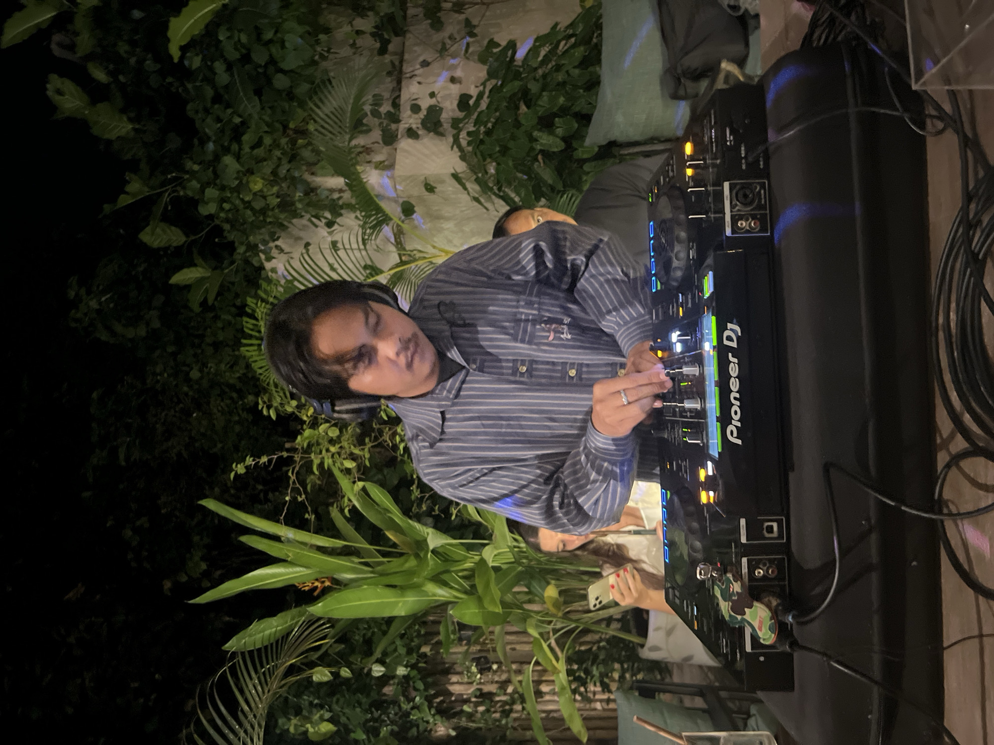 A full-shot, eye-level, outdoor evening photograph captures a male DJ with dark hair and a mustache, wearing a blue and white striped button-down shirt and headphones, engrossed in operating a Pioneer DJ controller. The DJ's hands are actively engaged with the equipment, with a silver ring visible on the left hand's ring finger. Behind the DJ, a woman with long dark hair, wearing a white top, is partially visible, looking at her mobile phone. To the right of the DJ, another individual, a man with short dark hair wearing a black t-shirt, is peeking from behind the DJ equipment. The background consists of lush, dense green foliage, suggesting an outdoor setting, possibly a garden or courtyard, with hints of a wall or structure behind the plants. The scene appears to be in the Ubud District, Indonesia, as indicated by the atmospheric lighting and tropical vegetation. The DJ equipment is illuminated, highlighting its buttons and screens, and various cables are visible in the foreground, suggesting an active music setup.